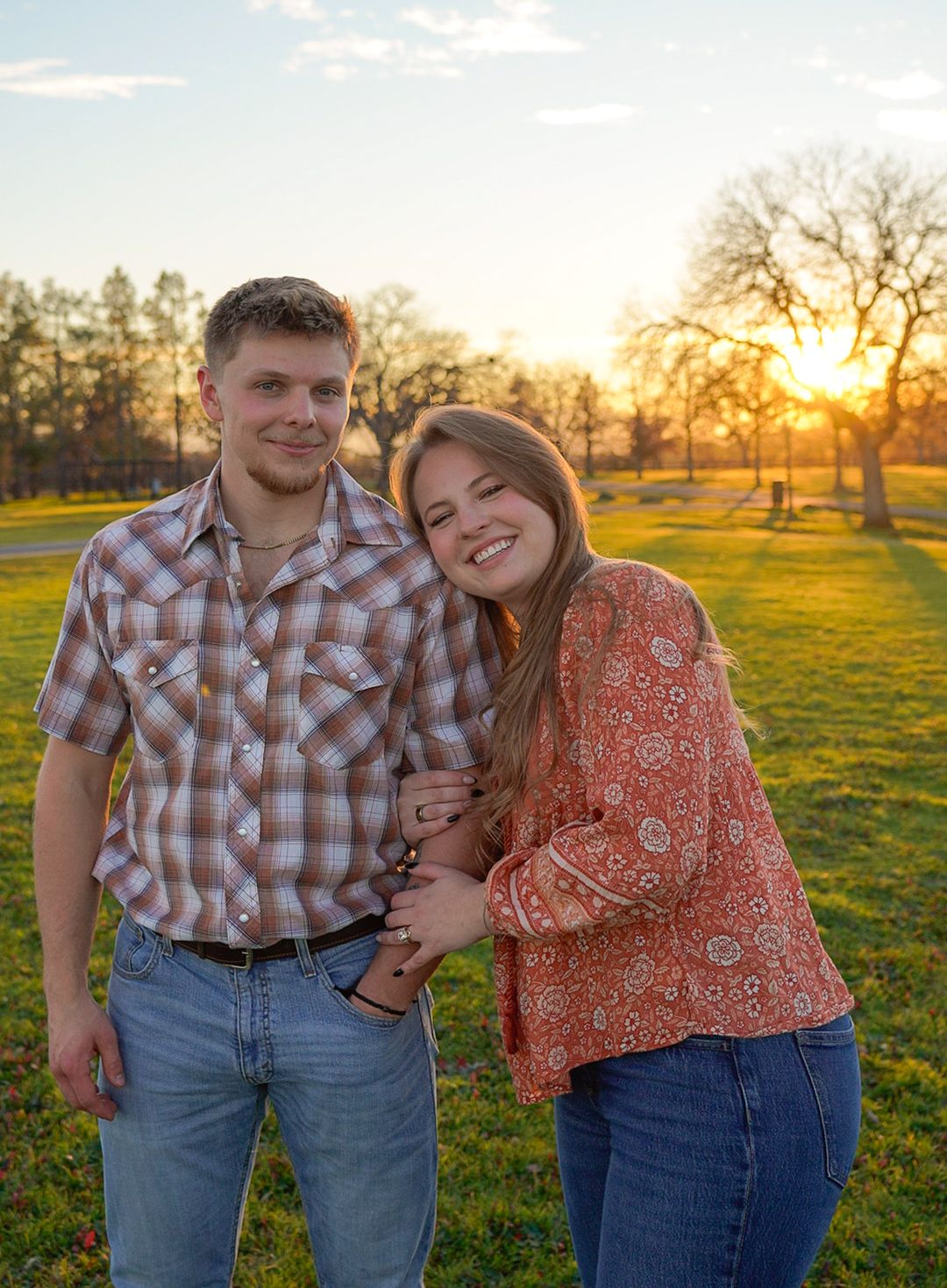 A young couple smiles together at golden hour in a park, man in plaid shirt and woman in floral top.