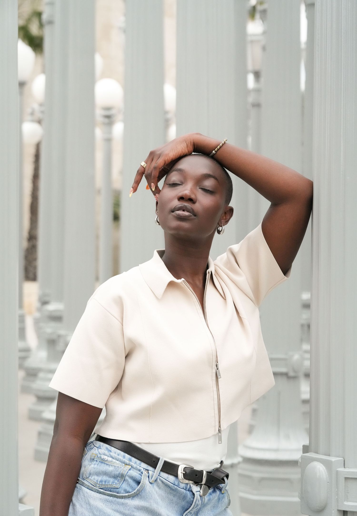 A young Black woman poses confidently in a cream zip shirt and jeans near white pillars, hand raised to her head.