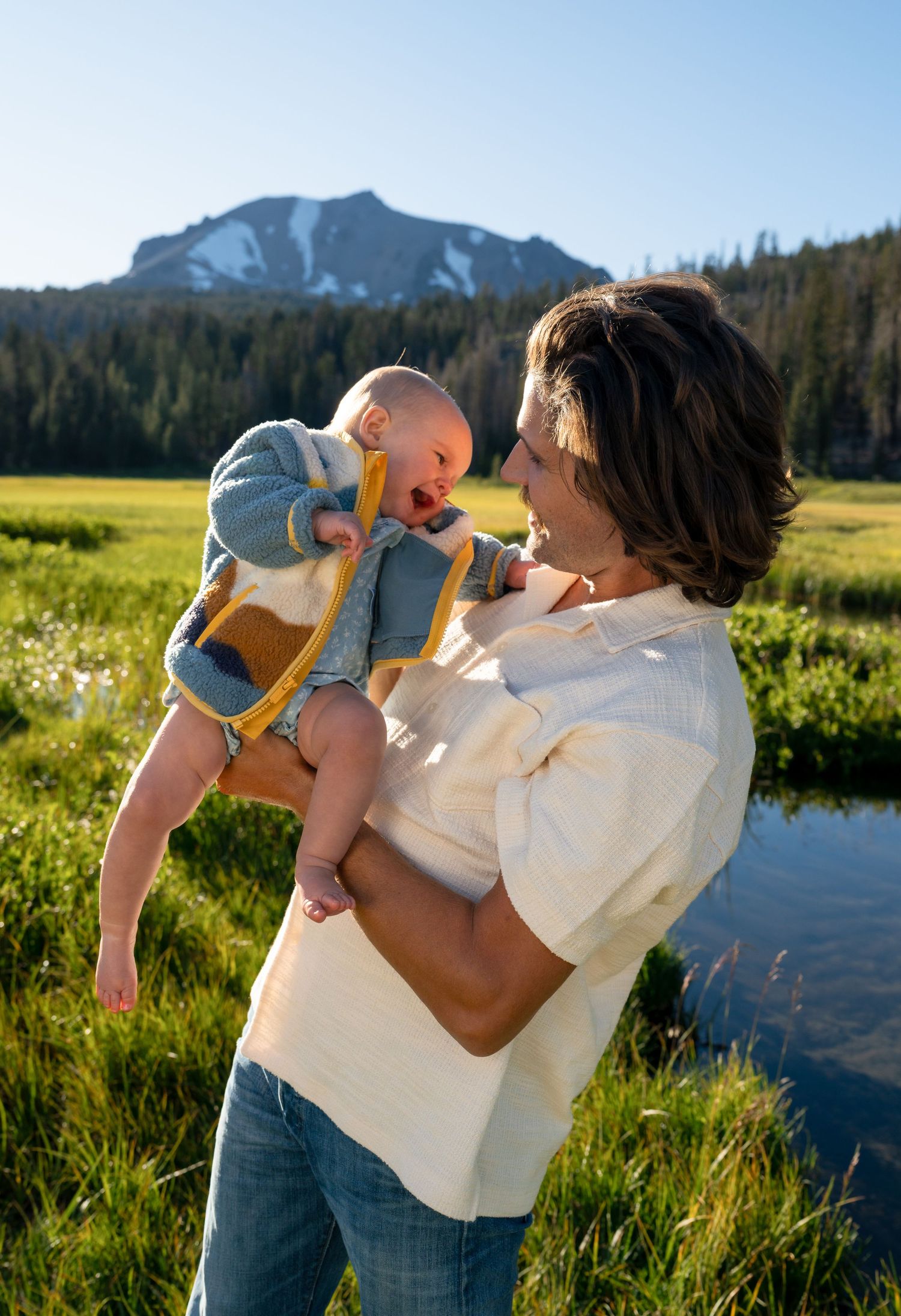 A parent holds a laughing baby outdoors in a green meadow with a snow-capped mountain and trees in the background.