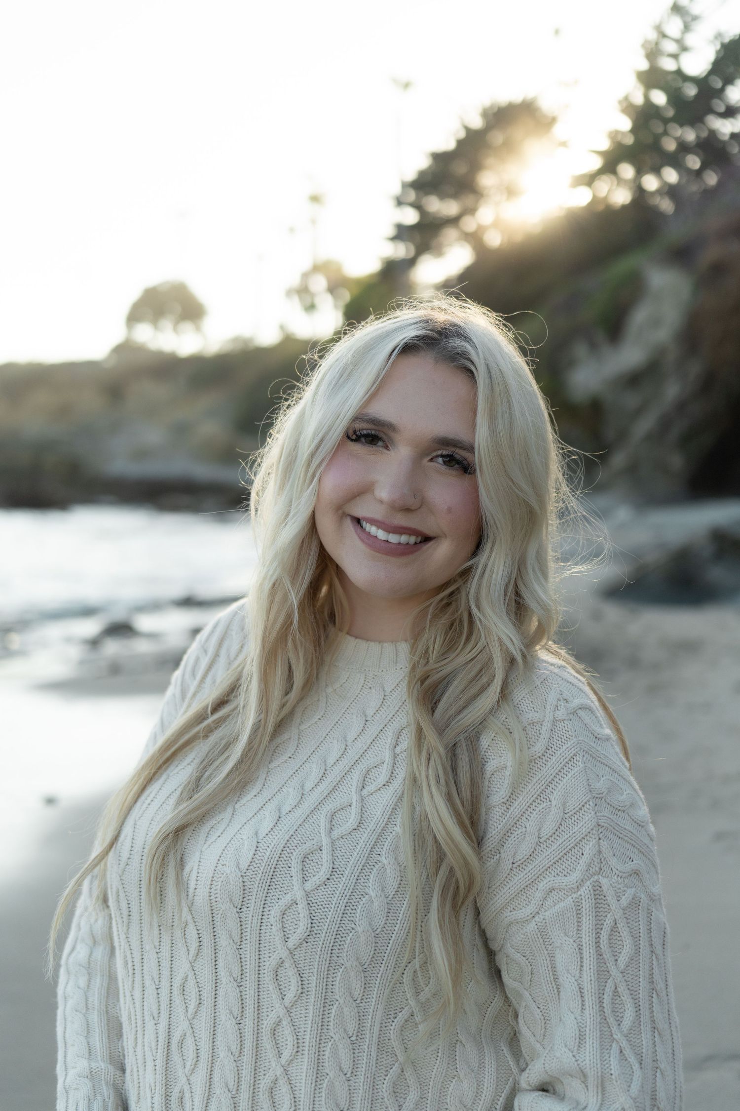 Smiling blonde woman in a cream cable-knit sweater poses on a beach at golden hour.