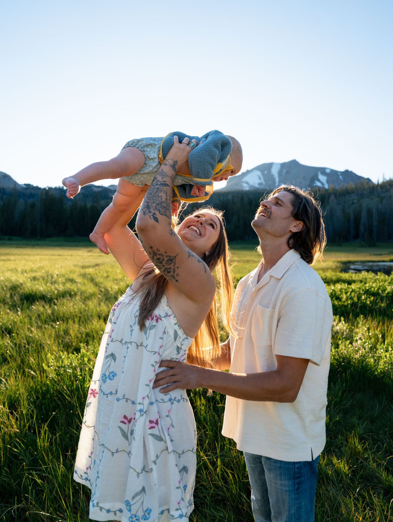 A joyful young family lifts their baby in a sunny mountain meadow, with green grass and peaks in the background.