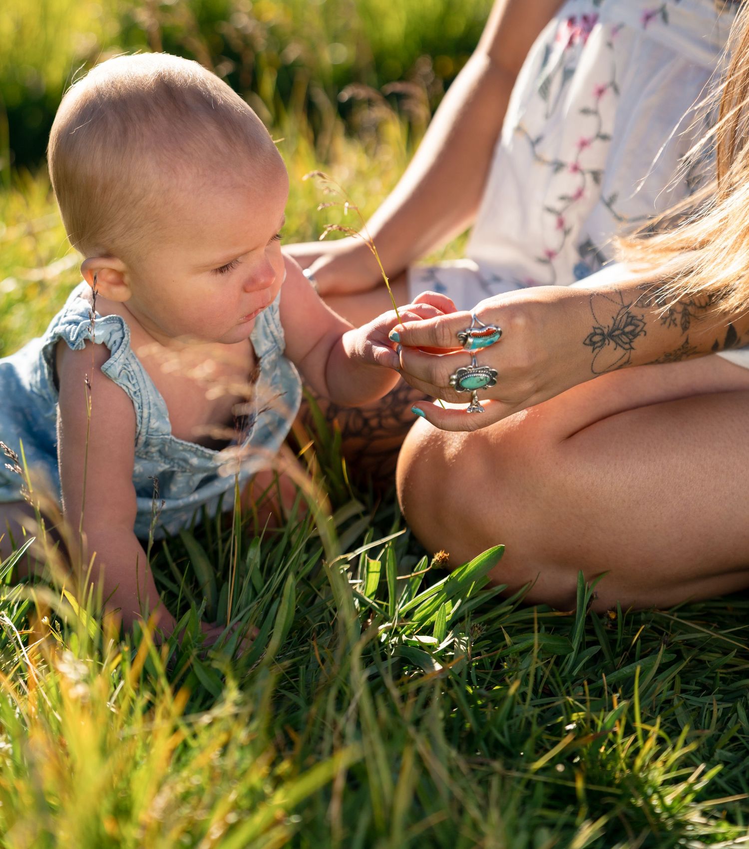 A tattooed mother sits in grass showing turquoise rings to her curious baby in a sunny outdoor setting.