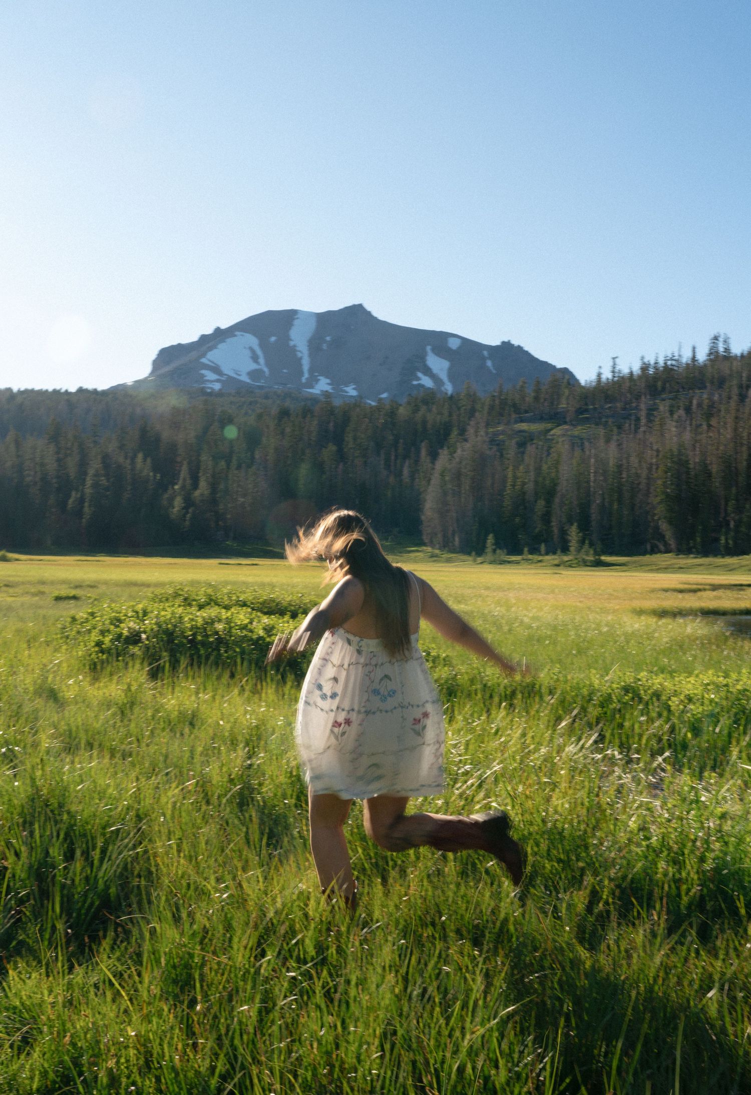 A girl in a white dress runs through a lush green meadow with a snow-capped mountain and pine trees behind her.