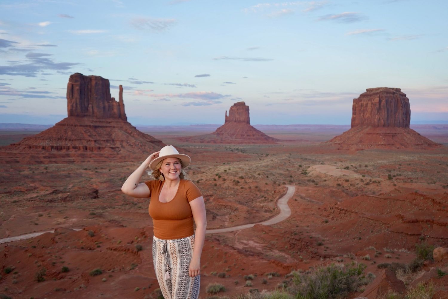 Woman in hat poses at Monument Valley with iconic red sandstone buttes and winding road in background.