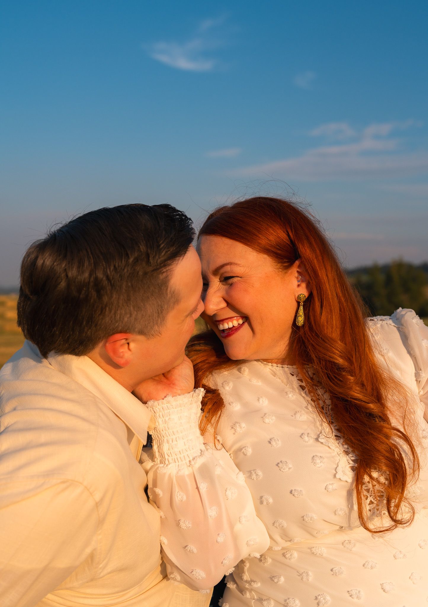 A joyful couple in white attire share a romantic moment outdoors during golden hour under a blue sky.