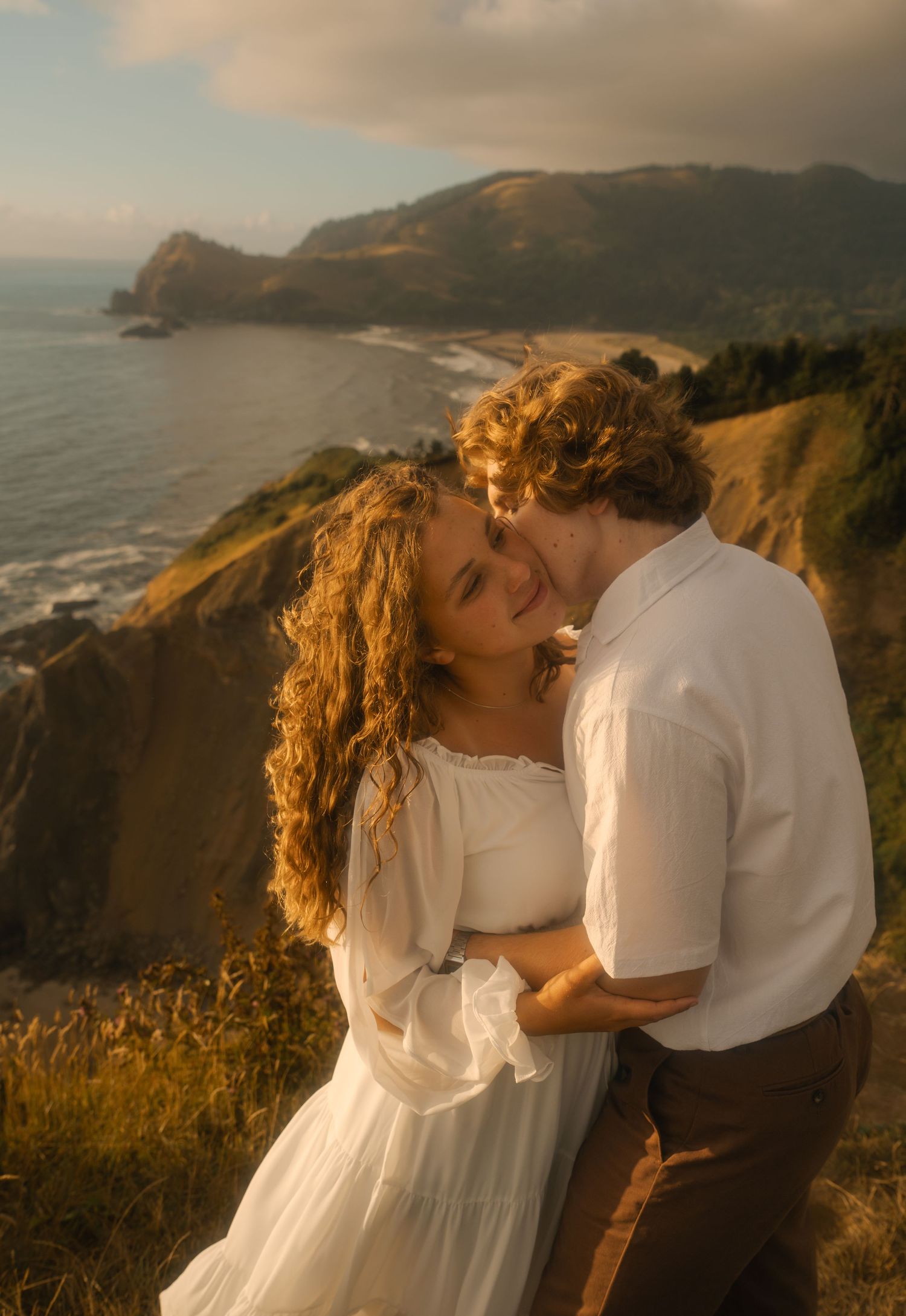 A couple shares a romantic kiss on a coastal cliff at golden hour, with ocean and mountains in the background.