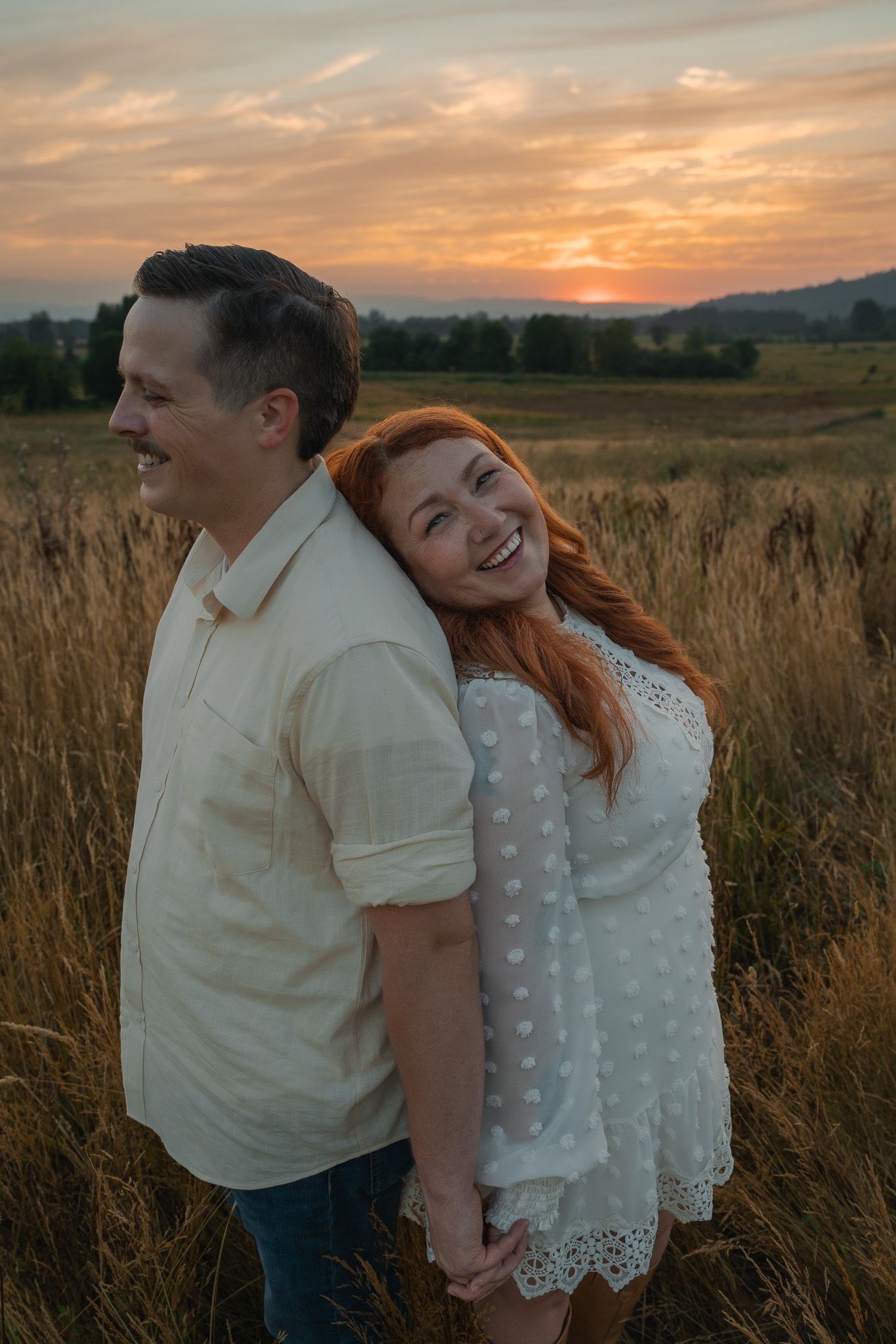Couple holding hands at sunset in a golden field, woman in white dress laughing, man in cream shirt smiling.