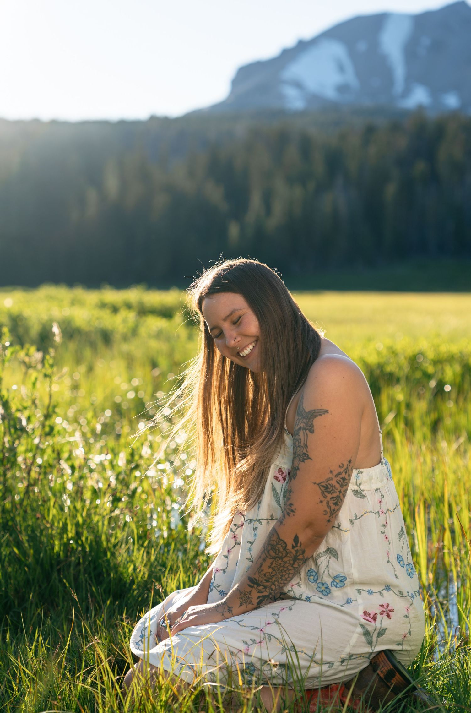 Smiling woman with tattoos in floral dress sitting in green meadow with mountain and forest backdrop.