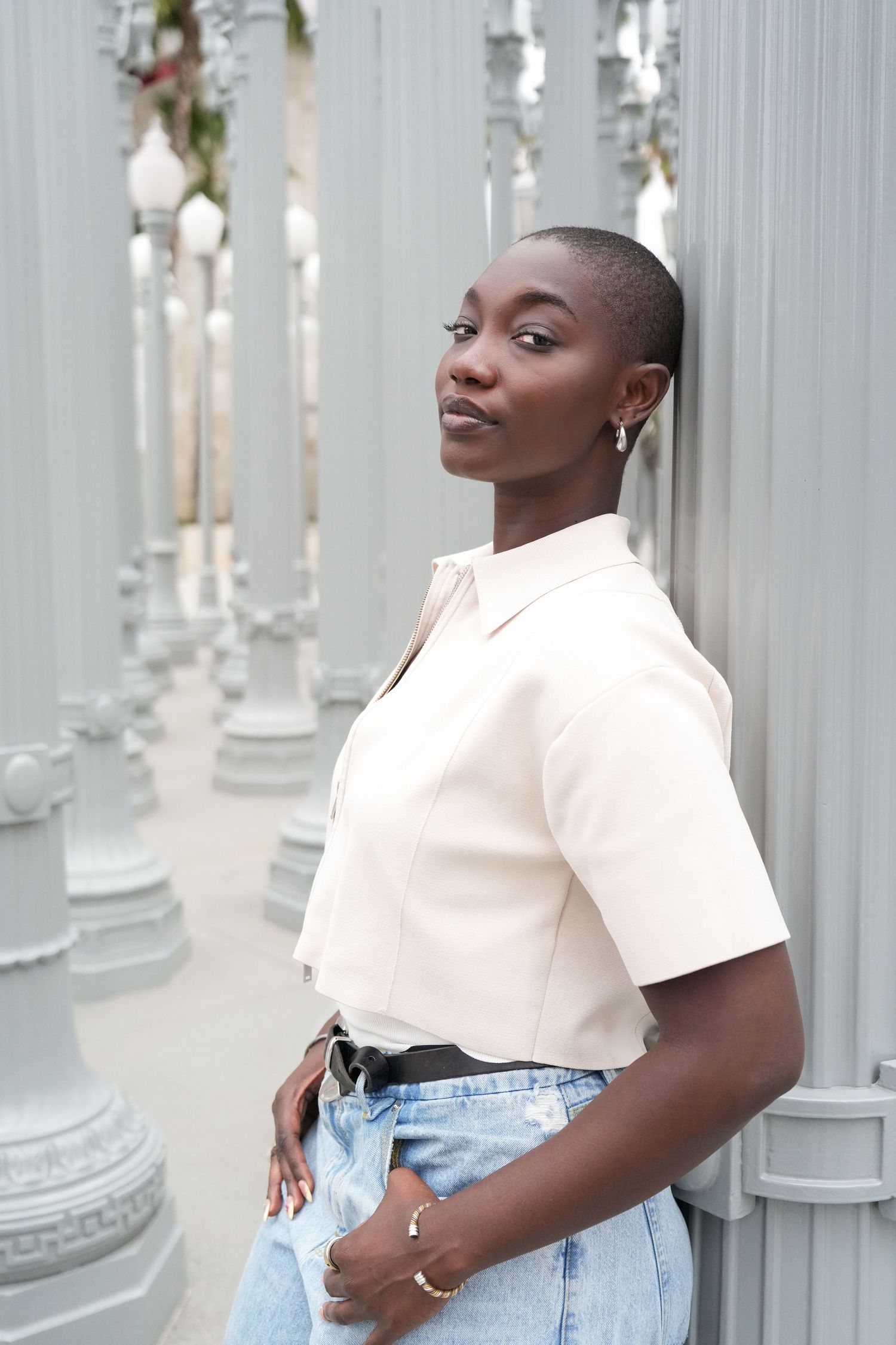 Young Black woman with shaved head in cream shirt and jeans leaning against gray pillars at LACMA.