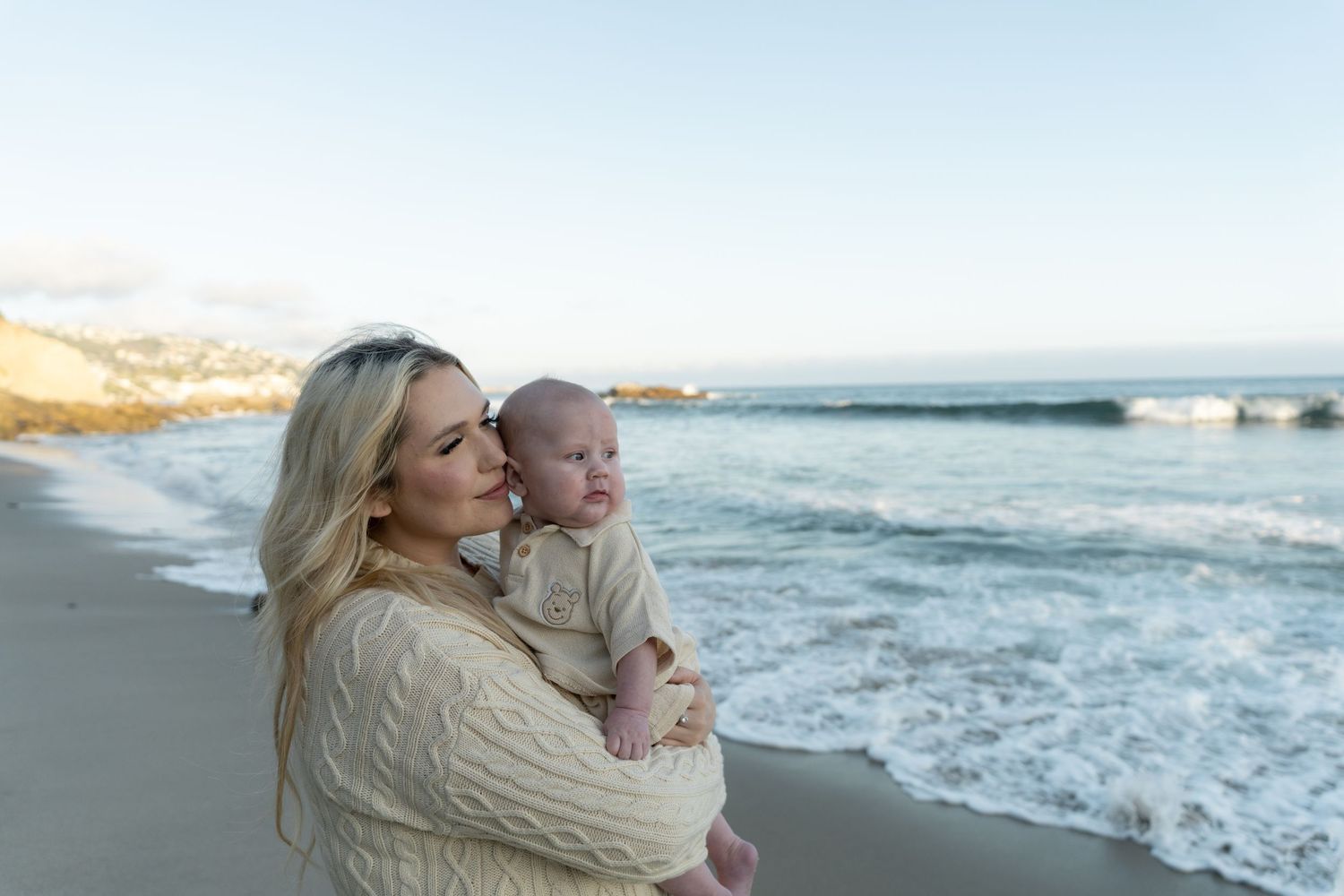 A blonde mother holds her baby on a sandy beach at sunset with ocean waves in the background.