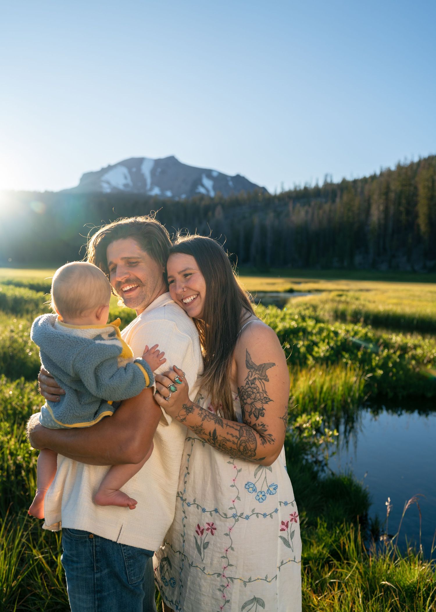 A smiling couple holding a baby in a sunlit mountain meadow with a peak and trees in the background.