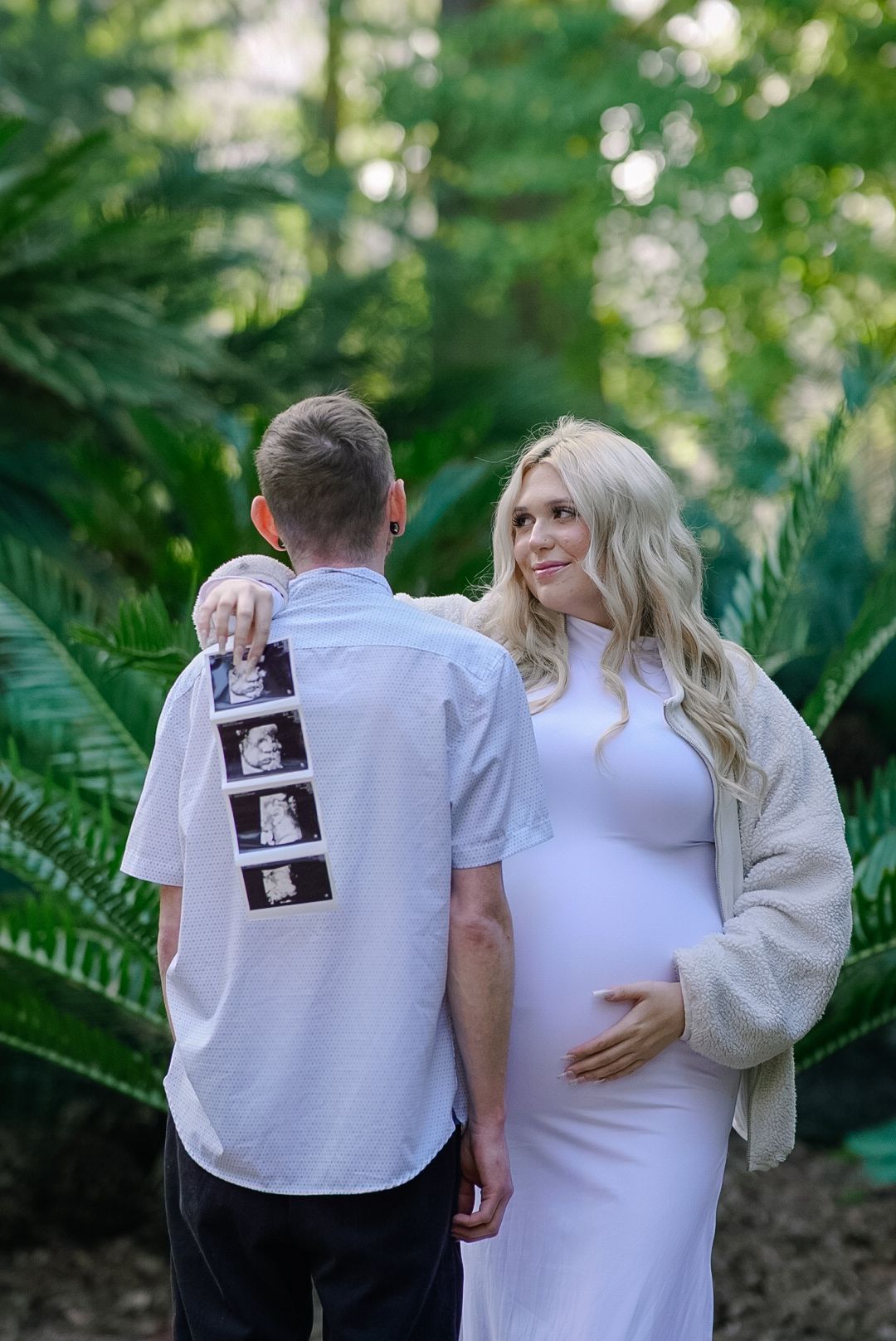 Pregnant woman in white dress holds baby bump beside partner showing ultrasound photos outdoors.