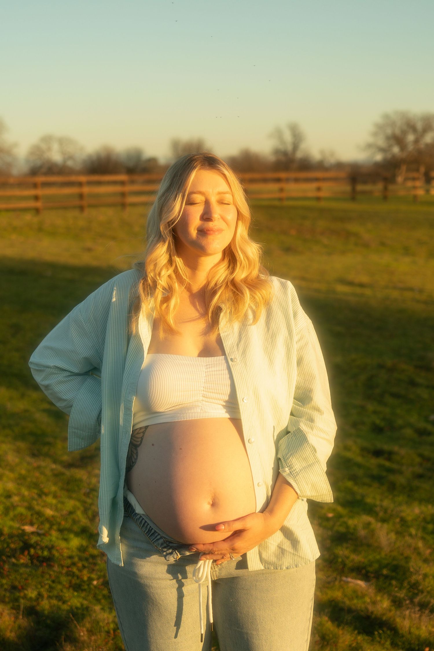 Pregnant woman in white crop top and denim jacket cradling her baby bump outdoors at golden hour near a wooden fence.