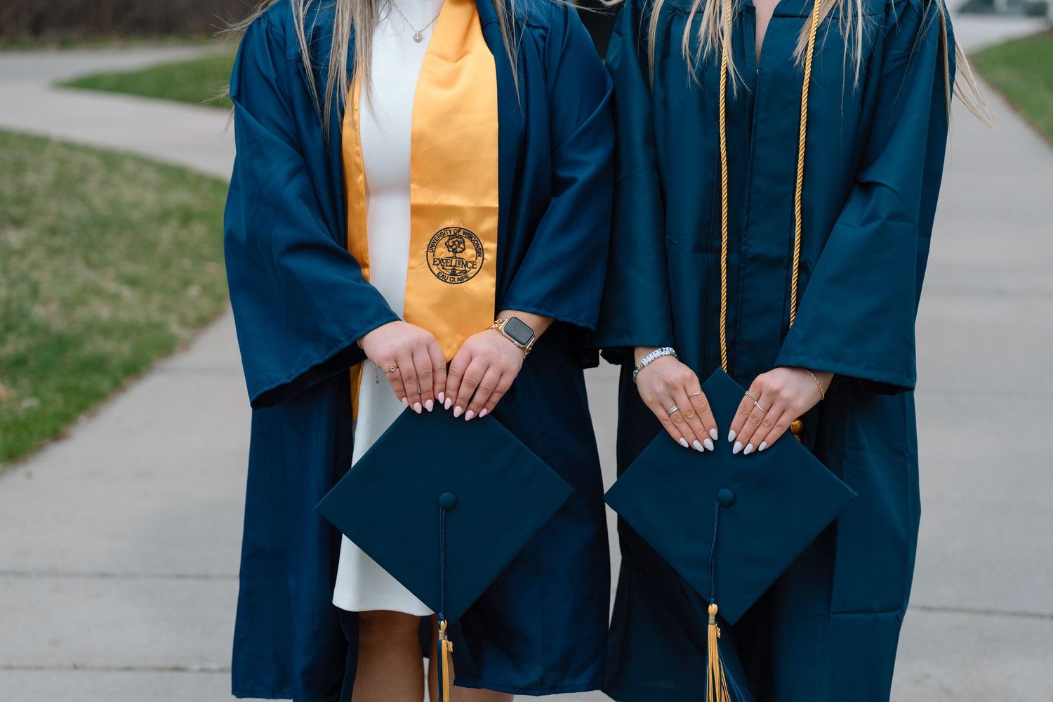 Two graduates in navy blue caps and gowns holding mortarboards, one wearing a gold honor stole, standing on a sidewalk.