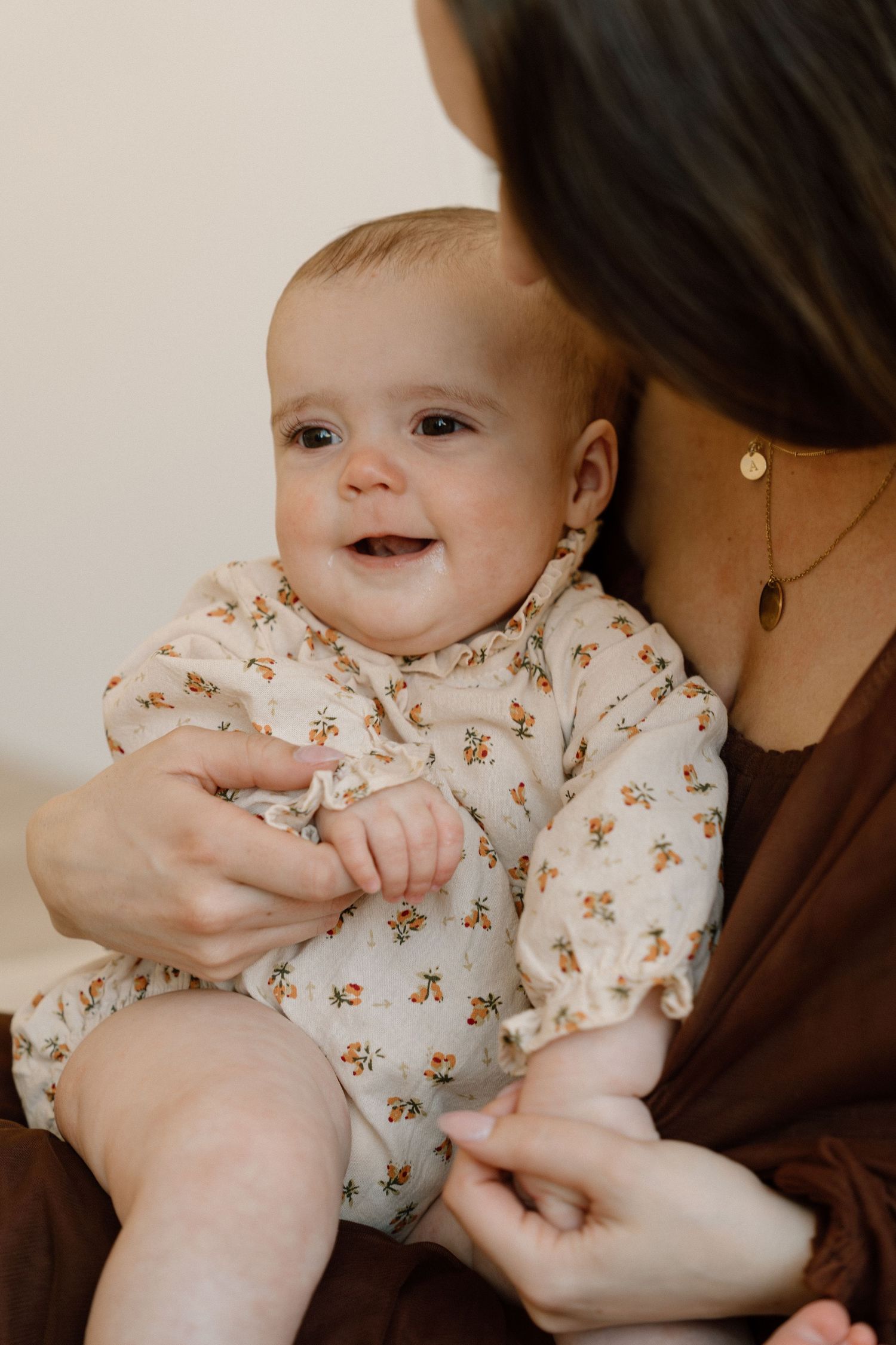 Smiling baby in floral romper being held by mother wearing brown dress and gold necklace.