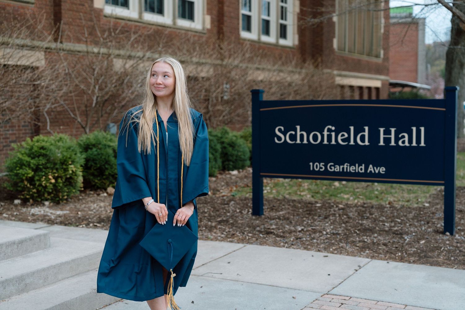 Graduate in teal cap and gown poses near Schofield Hall sign at 105 Garfield Ave on graduation day.