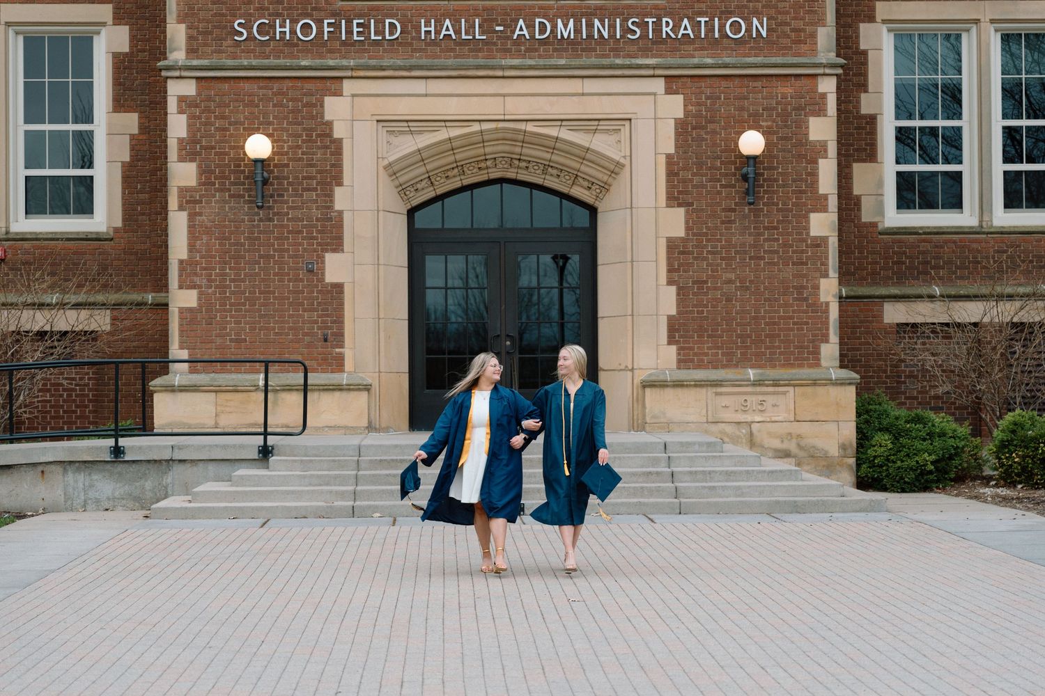 Two graduates in blue caps and gowns celebrate outside Schofield Hall Administration building on a college campus.