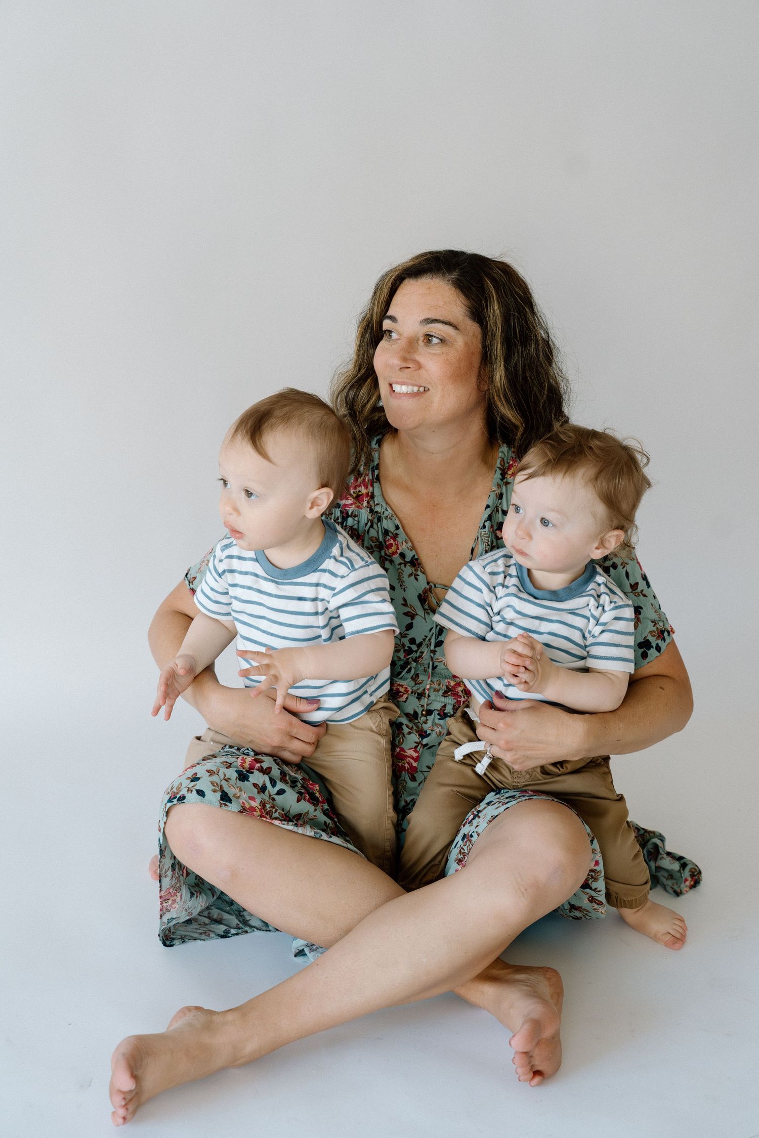 Smiling mother sitting cross-legged holding twin babies in matching striped shirts against white background.