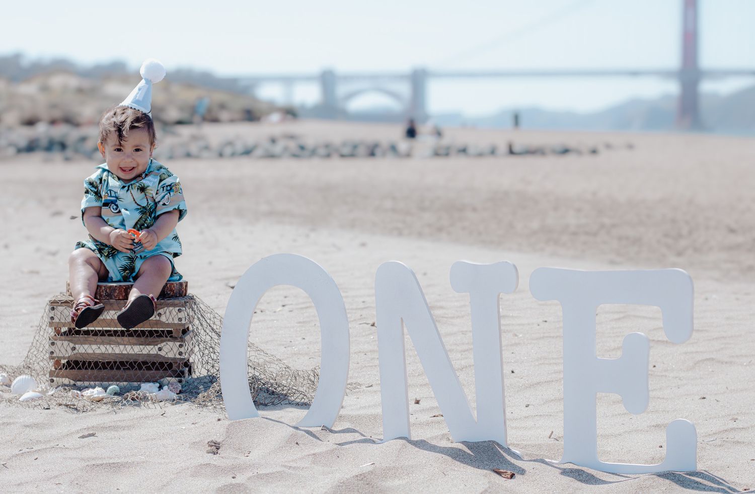 A small figure sits on a wicker basket beside large white 'ONE' letters on a sandy beach with a bridge in the background.