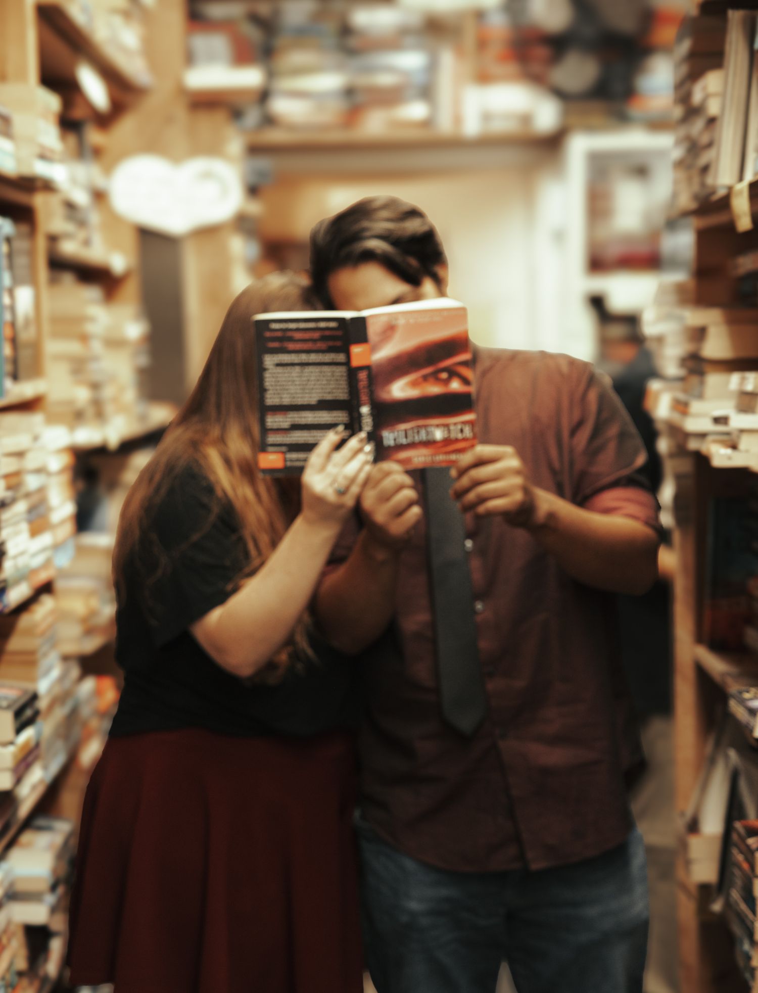 A couple holds books in front of their faces while posing in a cozy bookstore setting.