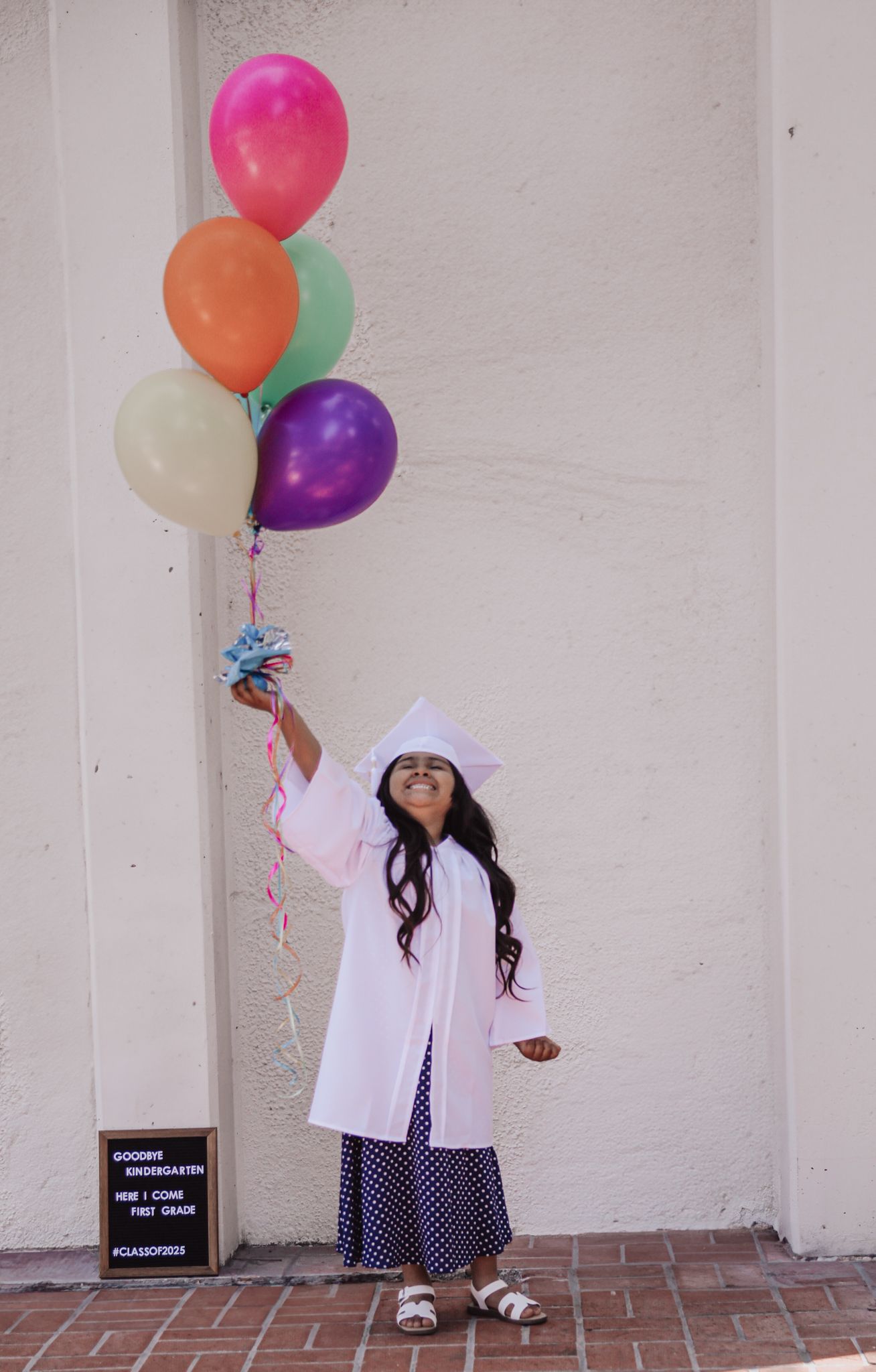 Young girl in white graduation cap and gown holds colorful balloons, celebrating kindergarten graduation outdoors.