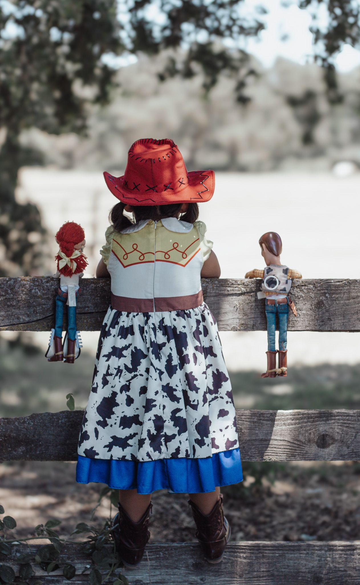 A young girl dressed as Jessie from Toy Story stands at a wooden fence with two Toy Story dolls beside her.