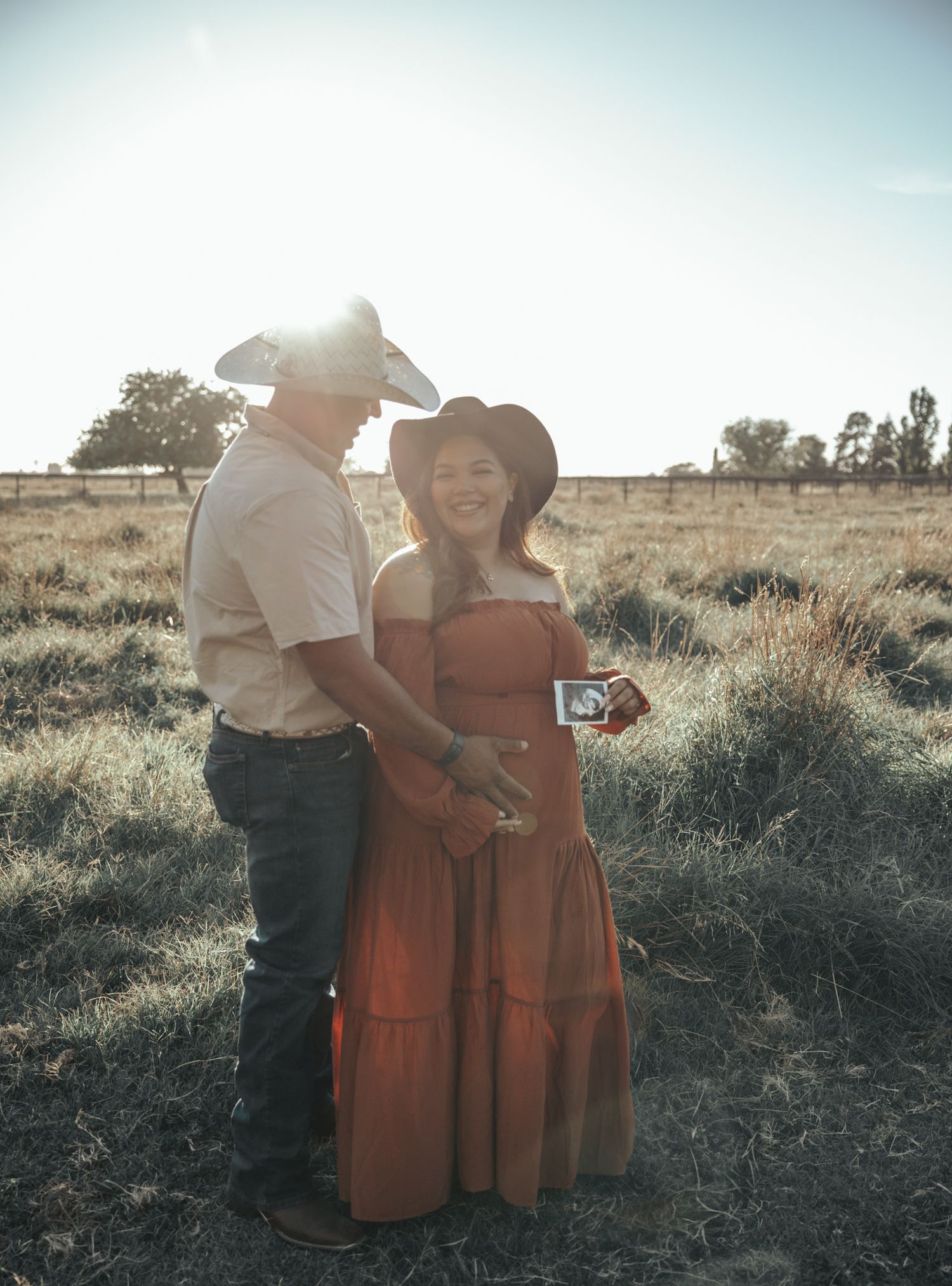 Couple in casual attire embraces while standing together in a sunlit field with dry grass and desert landscape.
