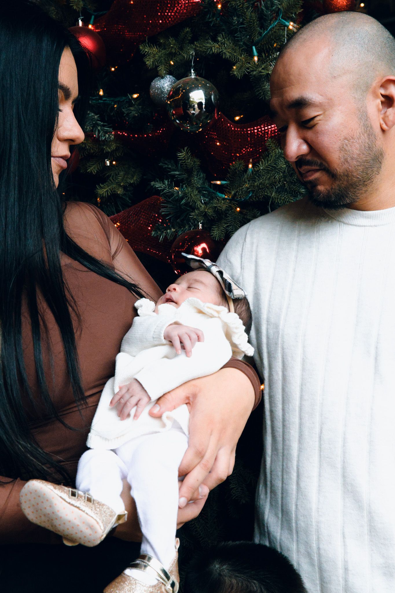 A couple holds and gazes lovingly at their newborn baby near a Christmas tree in a series of intimate photos.