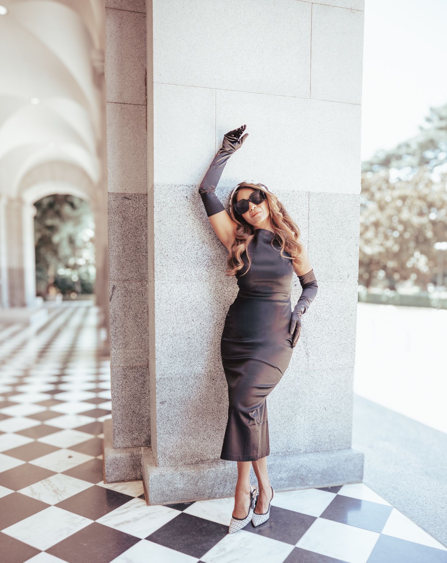 Woman in gray dress poses gracefully against a white column in architectural setting with checkered floor pattern.