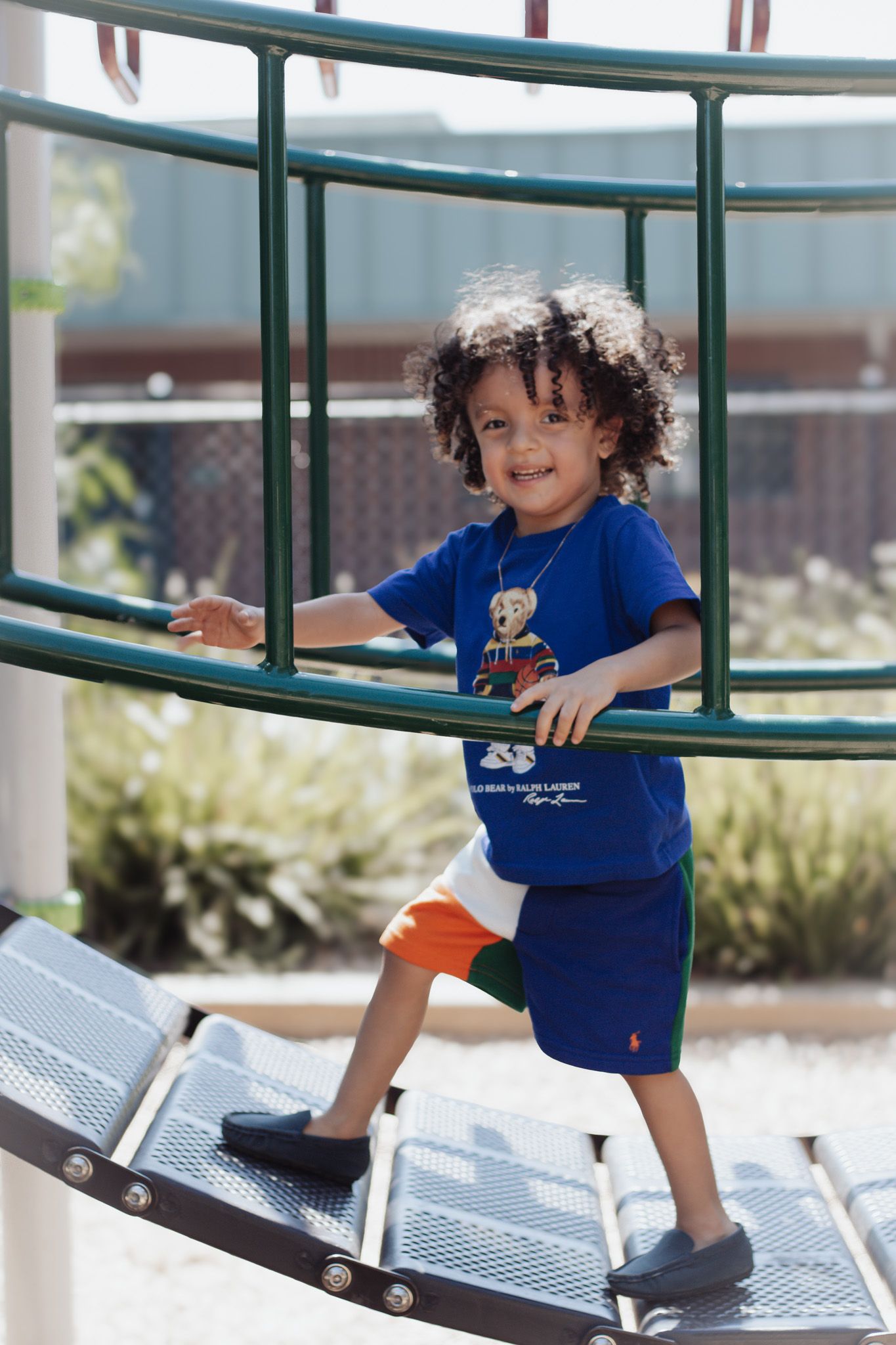 A small child wearing a blue shirt and shorts plays on outdoor playground equipment on a sunny day.