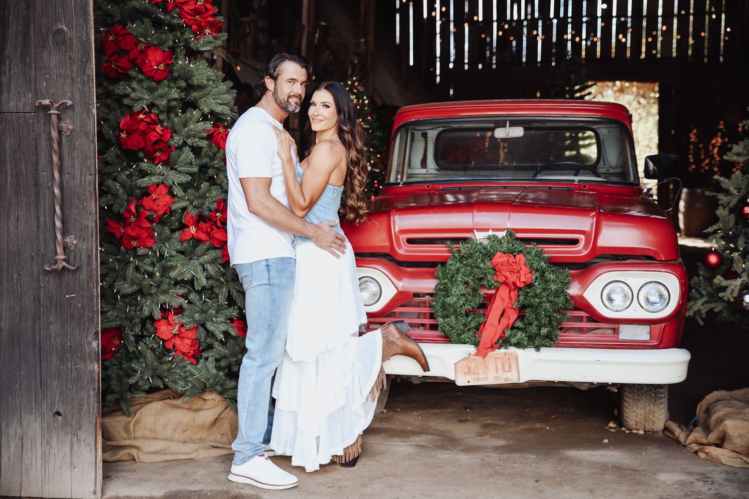 A couple poses in front of a vintage red Chevy truck decorated with a Christmas wreath and red roses in the background.