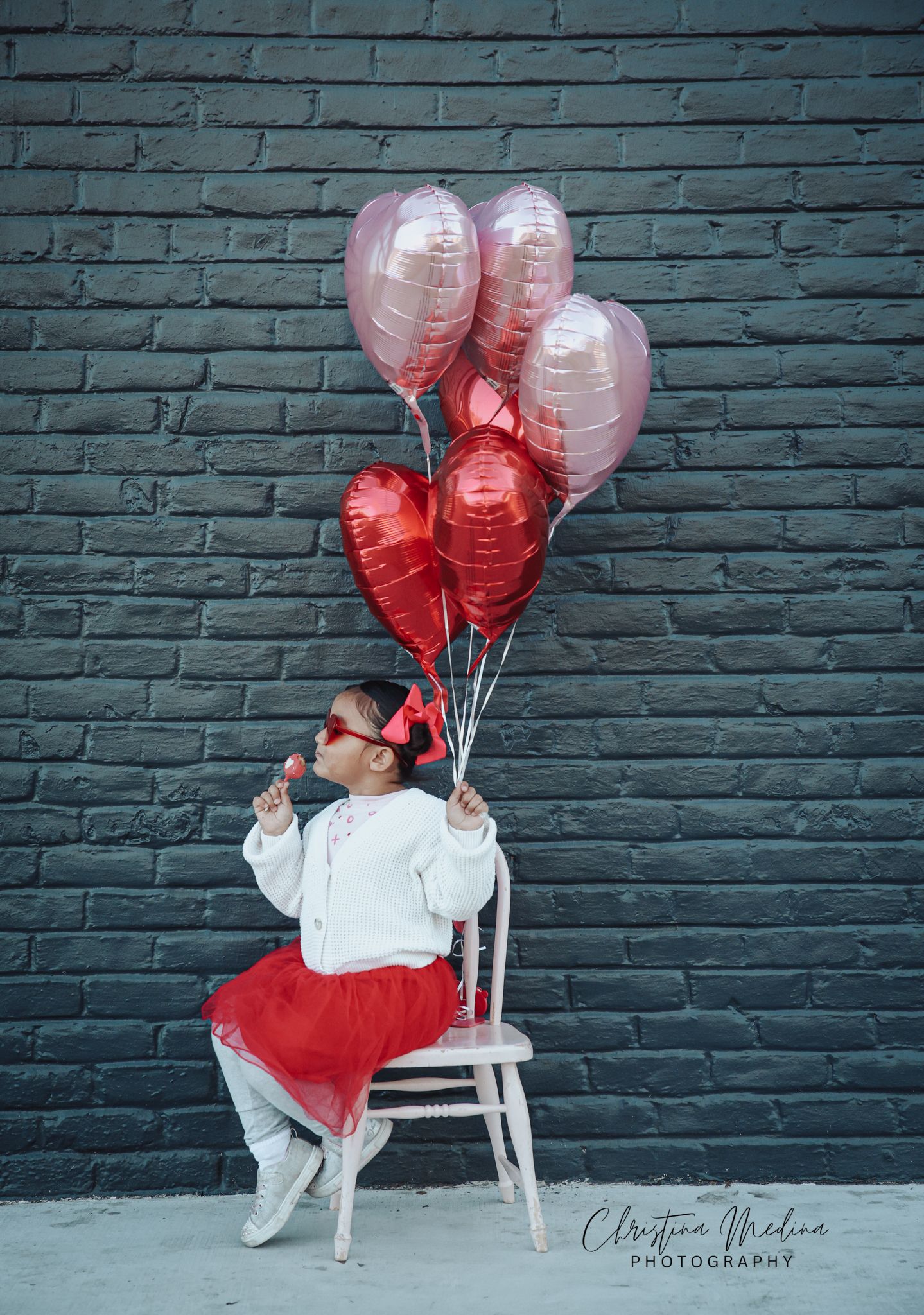 A child sits on a white chair against a teal brick wall holding pink and red heart-shaped balloons.