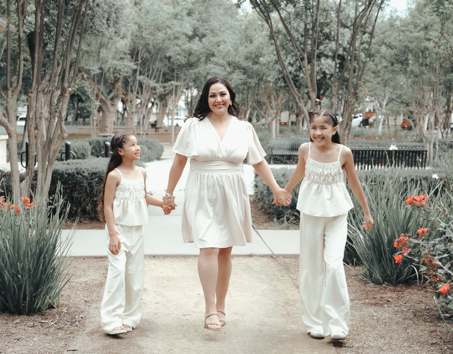 Three people wearing white outfits walk hand in hand along a garden path surrounded by trees and plants.