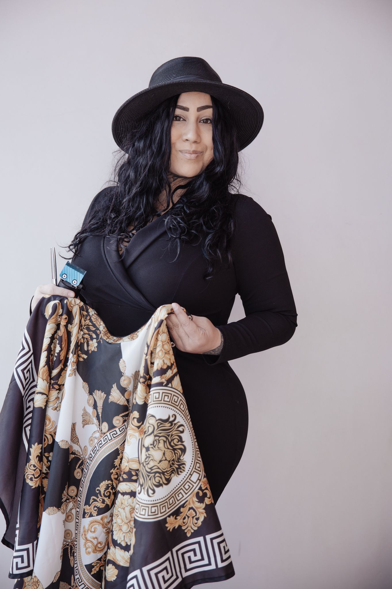 A model wearing a black outfit and wide-brimmed hat poses with a patterned scarf featuring Greek key designs.