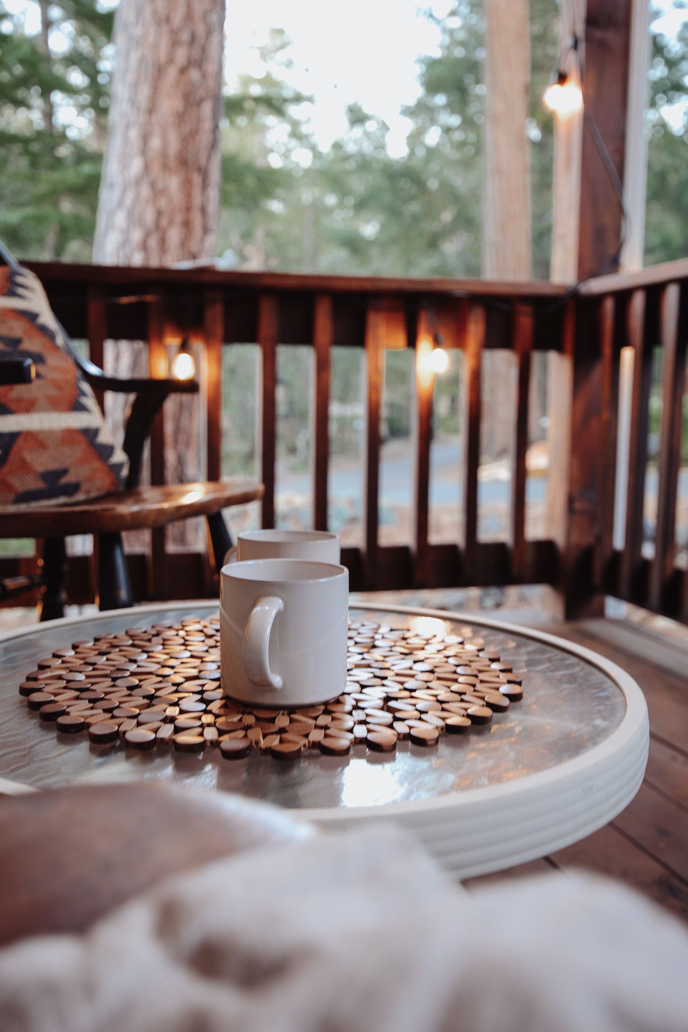 White ceramic mug on wooden coaster sits on rustic deck table with forest view and ambient lighting in background.