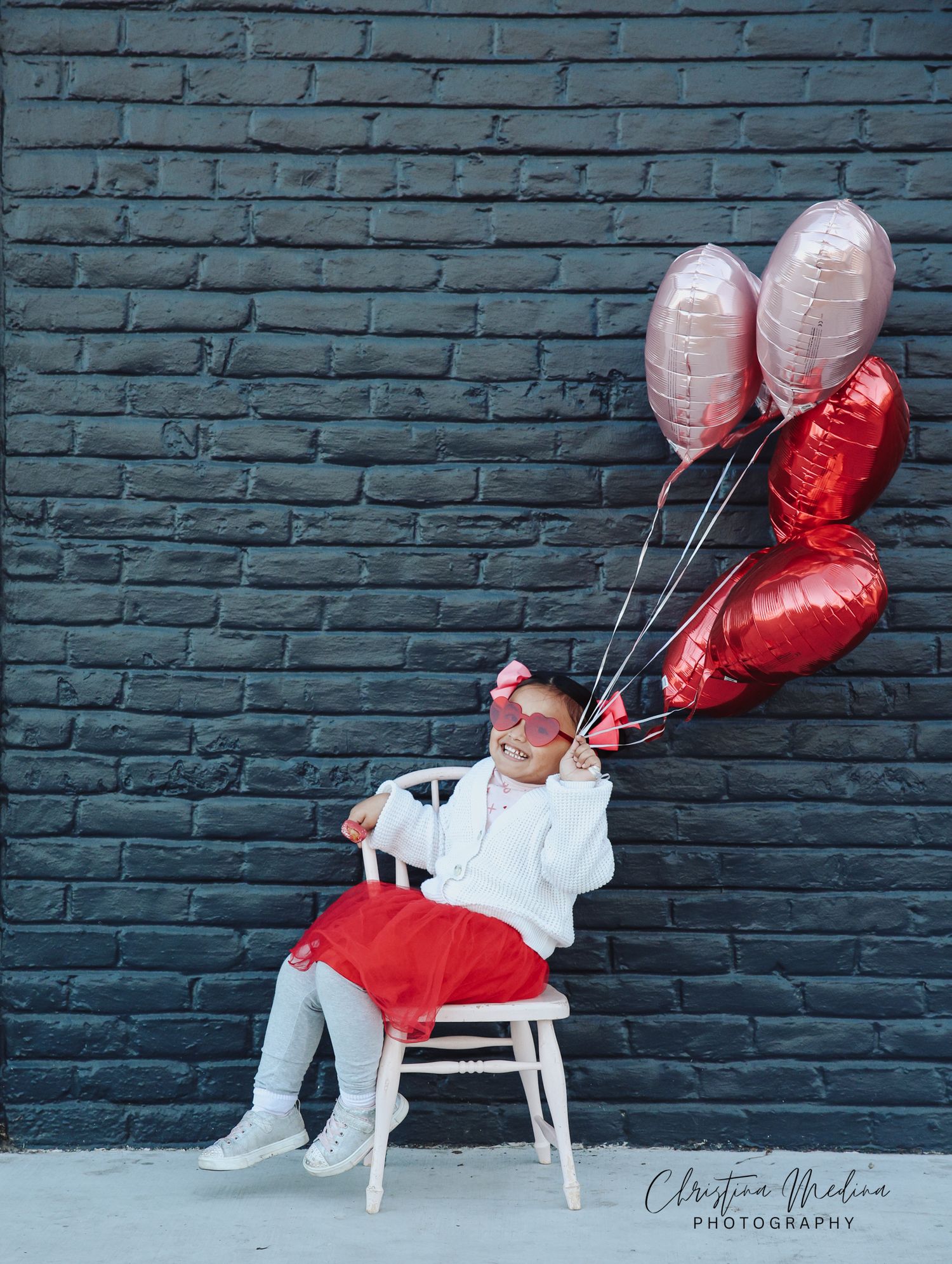 Baby in white outfit and red bow sleeps peacefully while floating with red heart balloons against grey brick wall.