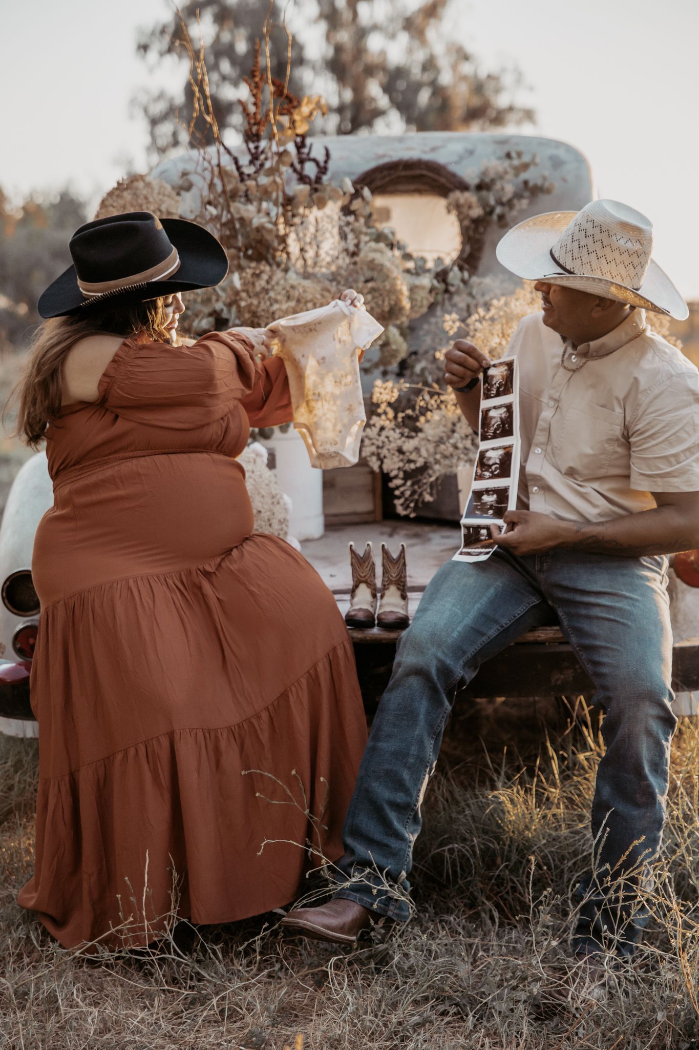 A couple in western attire sharing pregnancy announcement photos while sitting outdoors at sunset.