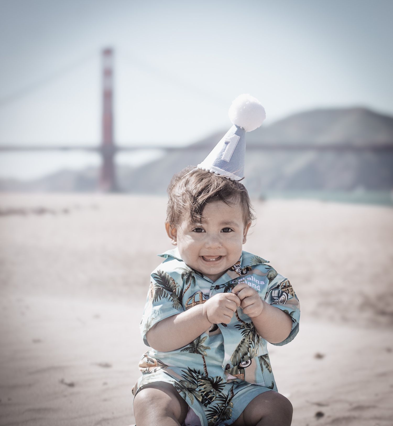 Child in party hat and floral shirt sitting on beach with Golden Gate Bridge visible in background on foggy day.