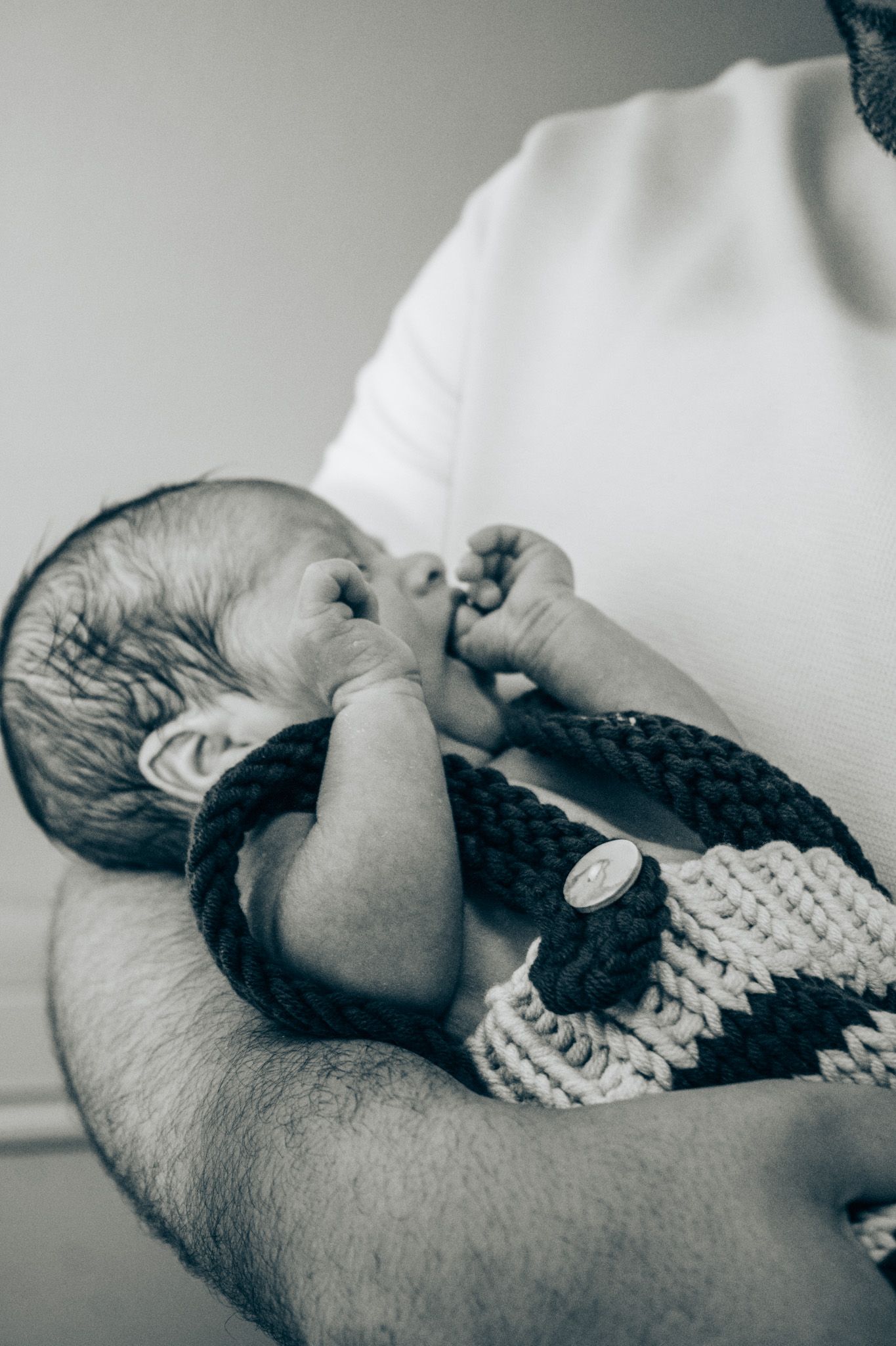 A newborn in a knit blanket sucking their thumb while being cradled in someone's arms.