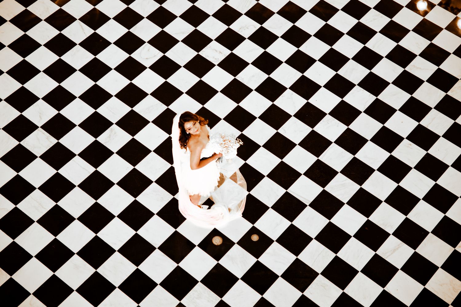 A couple in white attire embraces while standing on a dramatic black and white checkered floor pattern.