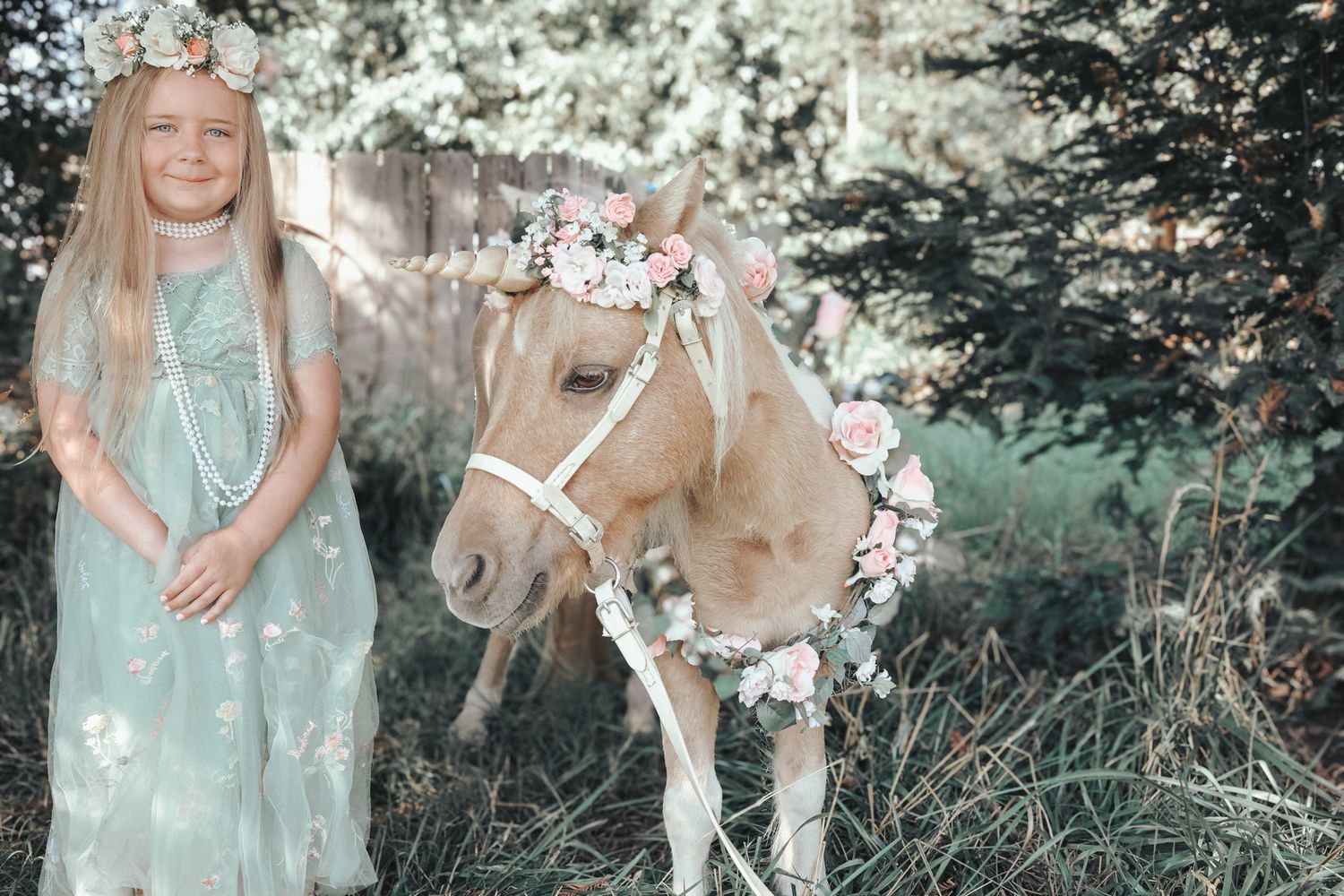Young girl in mint dress poses with a flower-crowned pony in a sunlit garden setting.