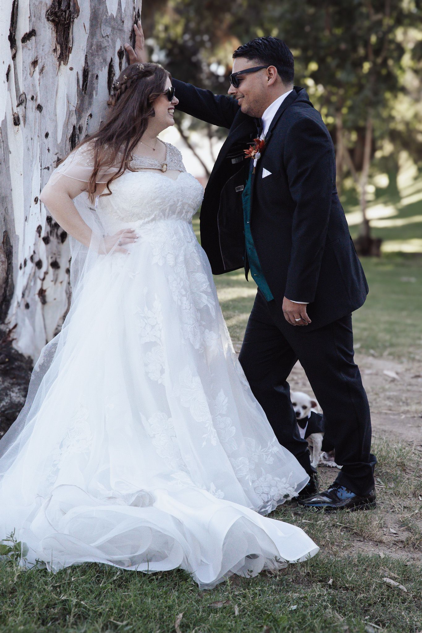 A bride in a white wedding dress and groom in a black suit share a tender moment under a tree in a park setting.