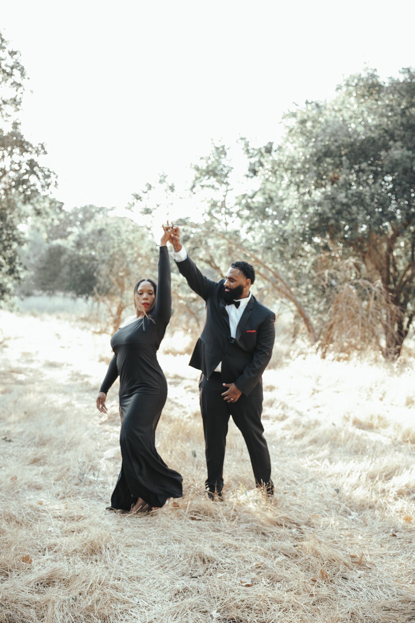 A couple in formal attire dances joyfully together on a hillside path surrounded by trees in soft natural light.