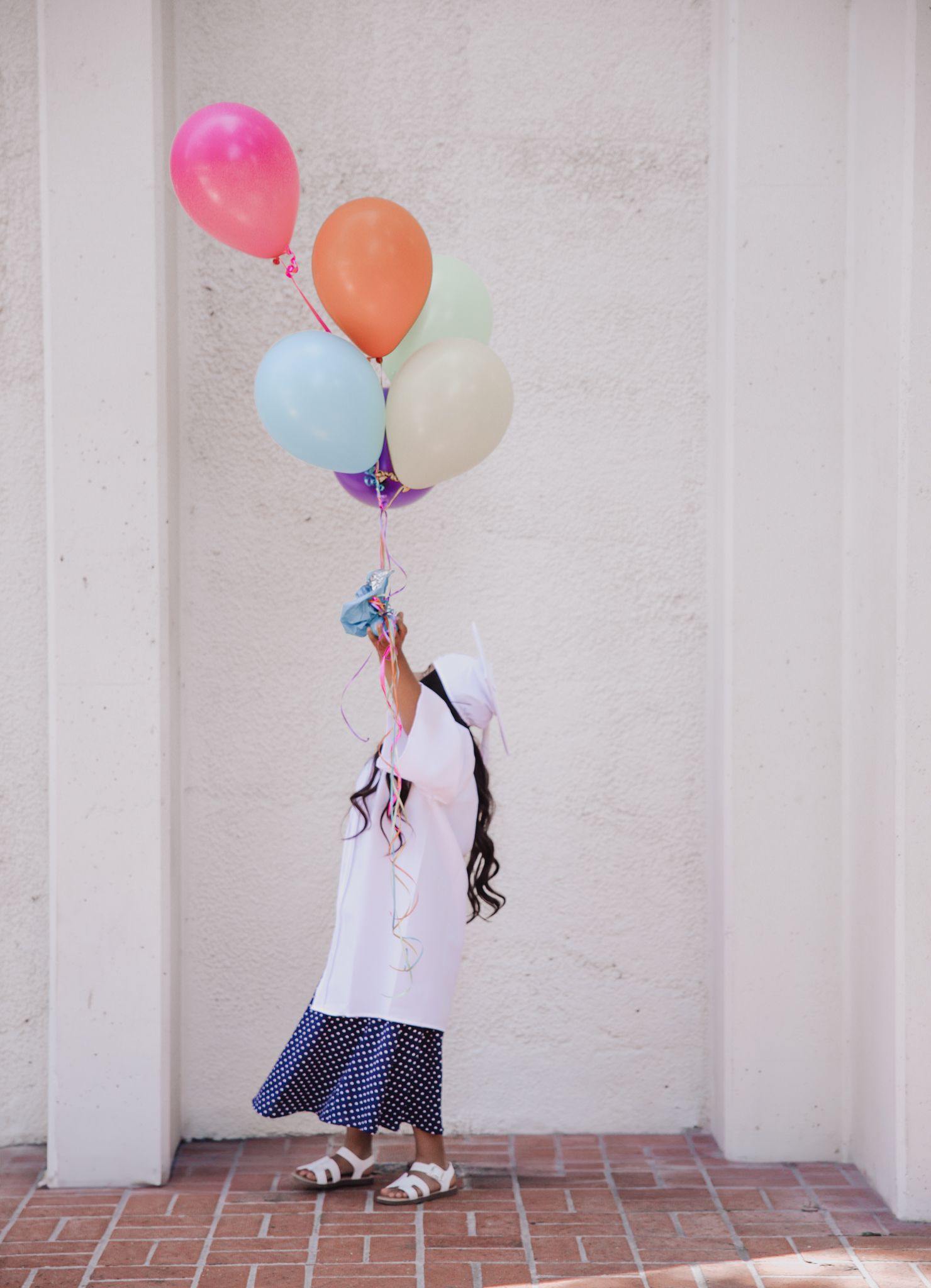 Girl in white dress holding colorful balloons against a white wall on a brick floor.