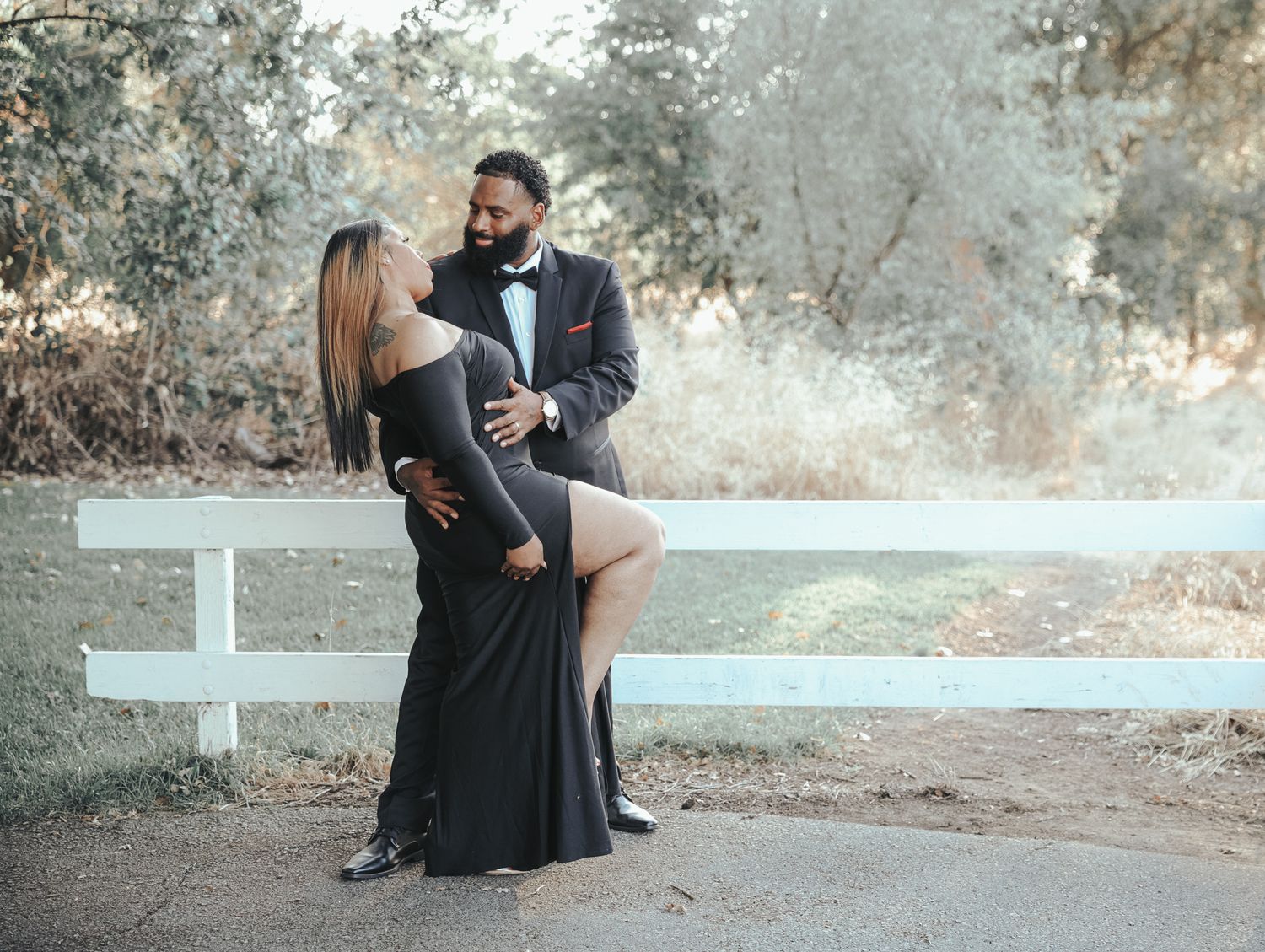 A romantic couple in black formal attire sits together on a rustic outdoor path during a photoshoot.
