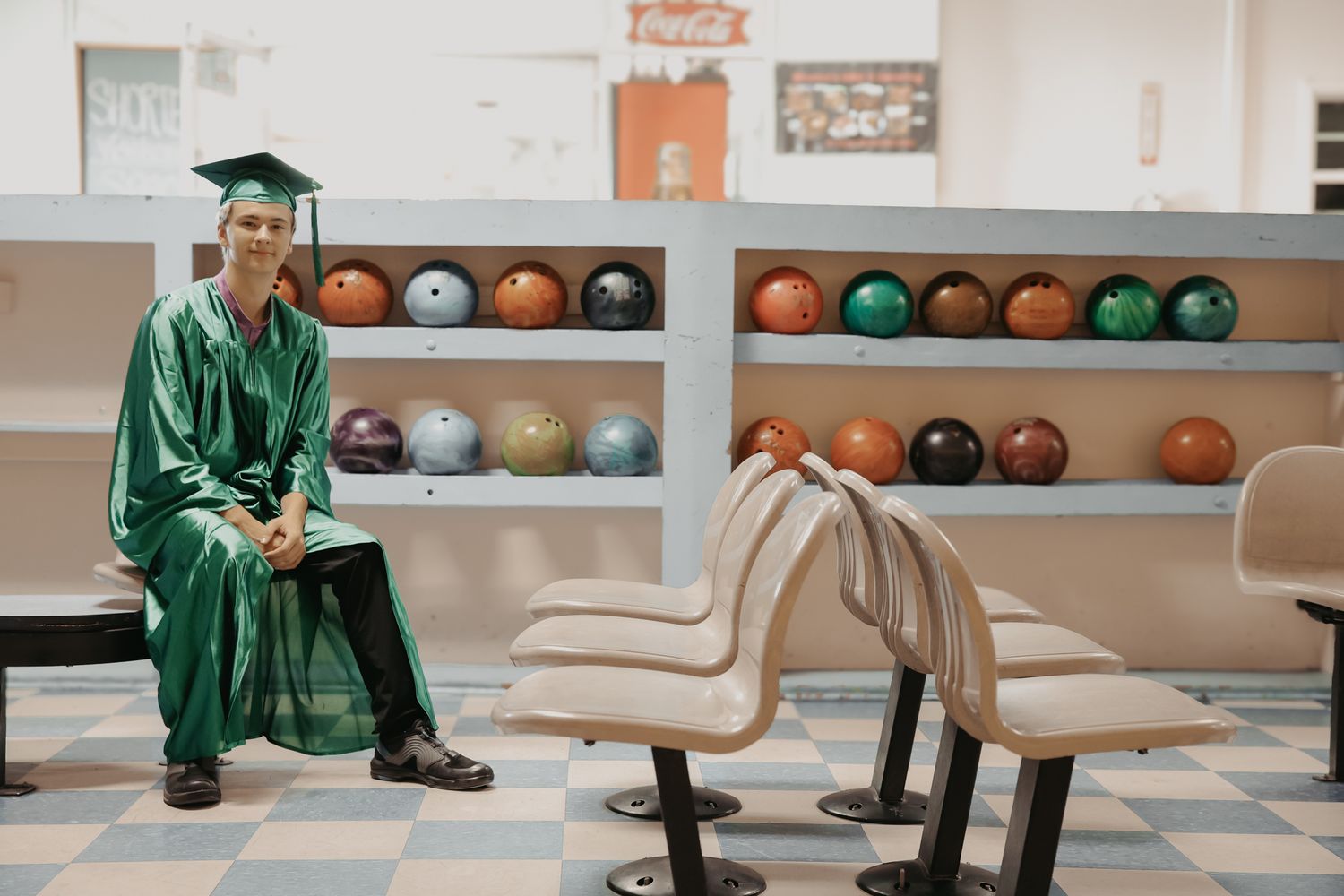 Graduate in green cap and gown sits on a wooden bench in a bowling alley with rows of bowling balls on shelves.