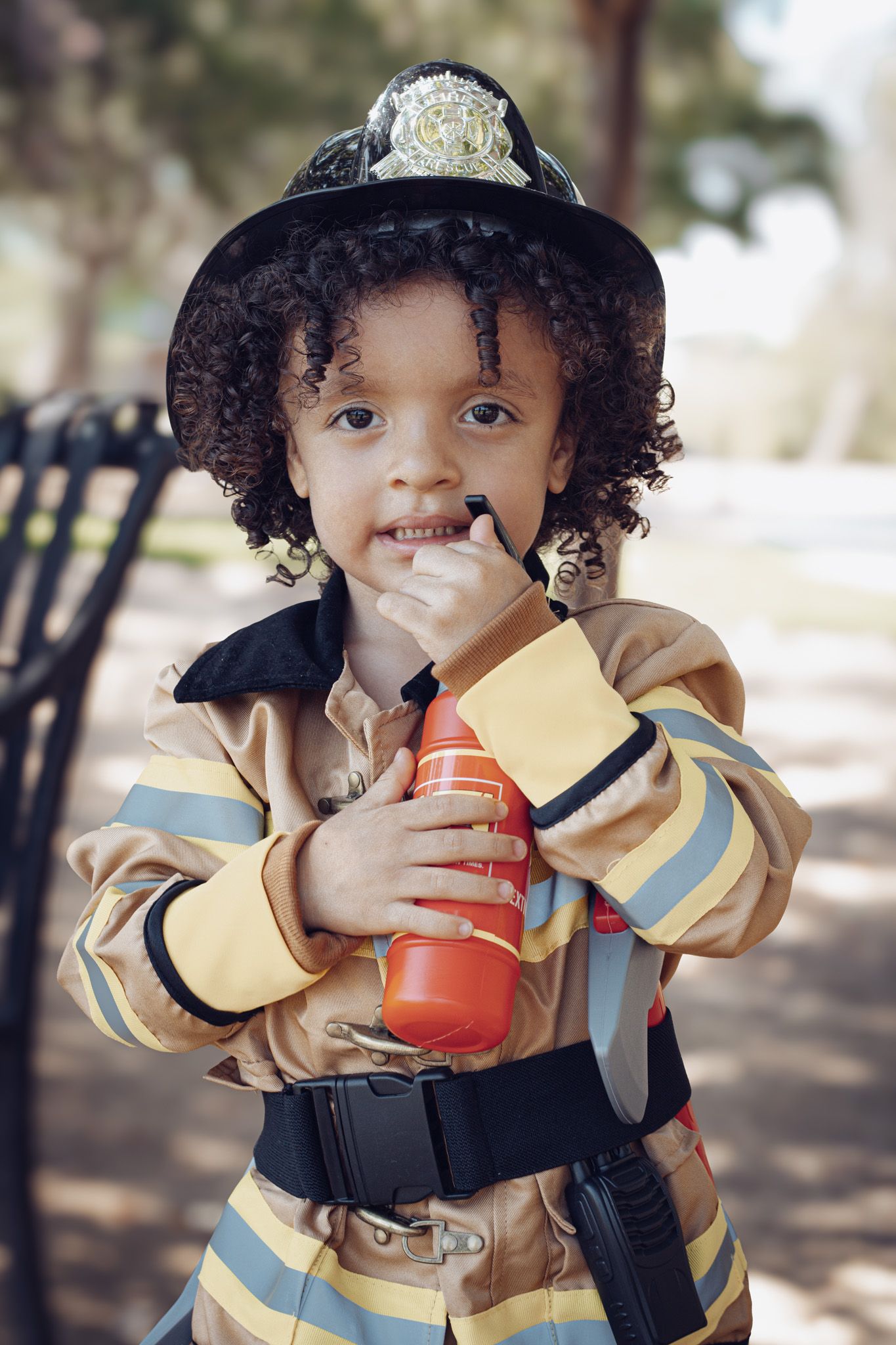 Young child wearing a firefighter costume with helmet and uniform holds a red bottle at a park.
