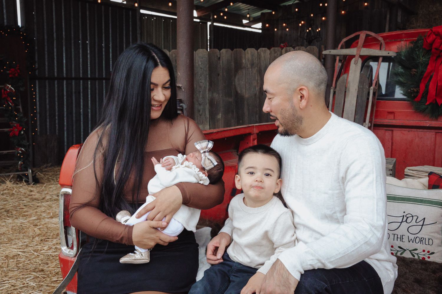 A family sits together outdoors near a red vehicle while holding a baby in white clothing.