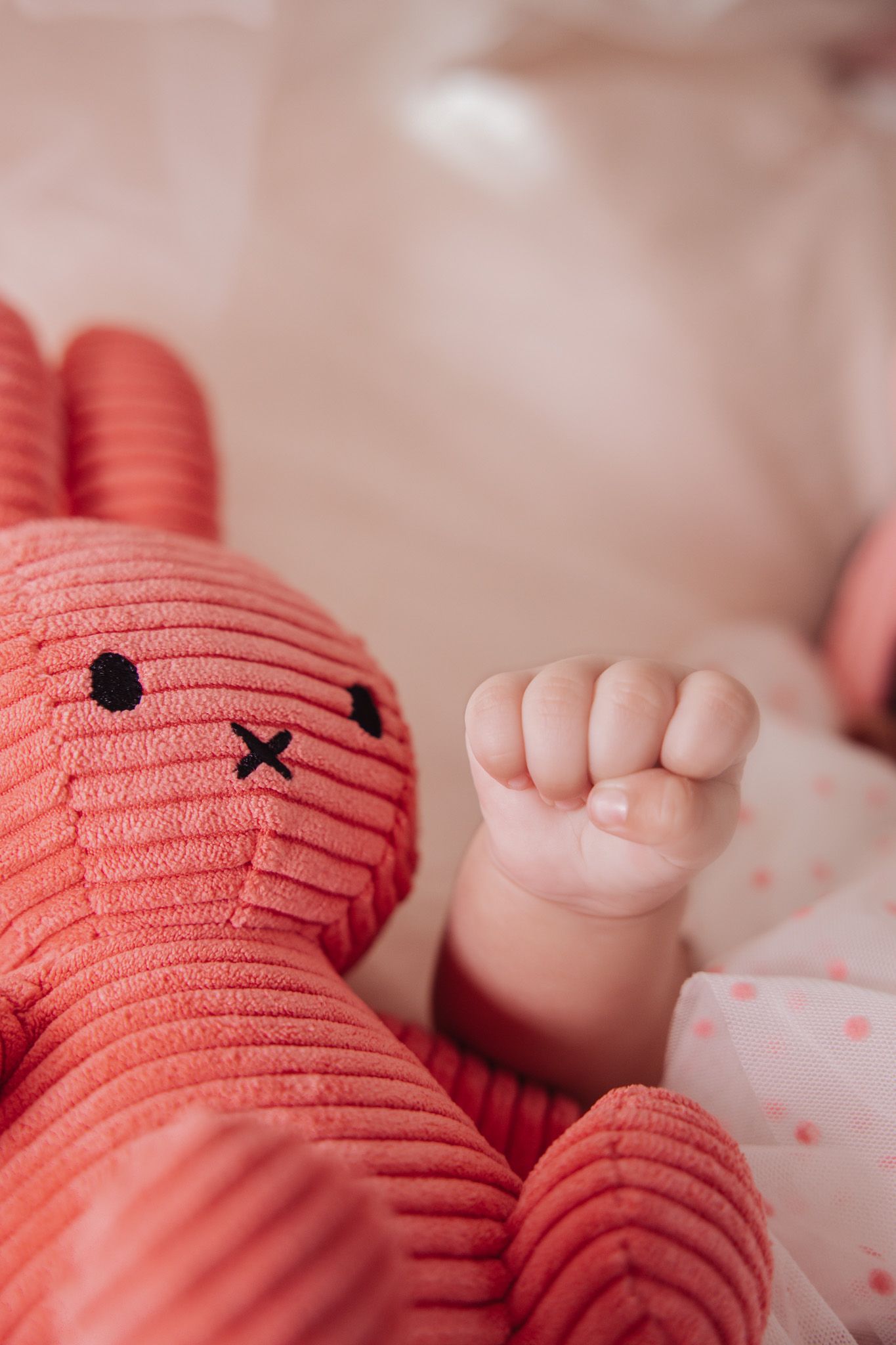 A tiny baby's hand rests next to a coral-colored plush bunny toy with embroidered facial features.