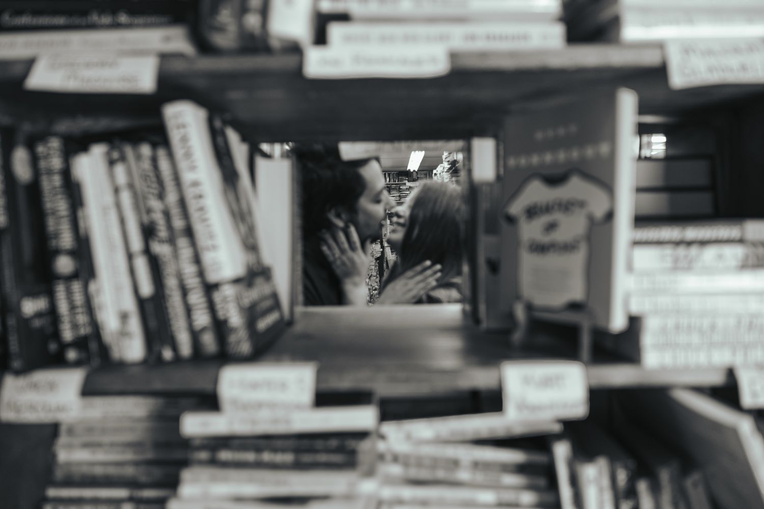 A black and white photograph focused through books on a shelf shows a figure holding a glass in celebration.